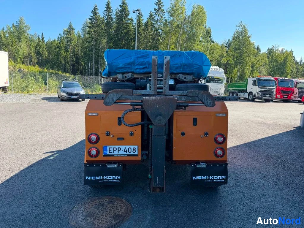 Takaa: Hina-auton takanäkymä: oranssit säilytyskaapit, kuorma sinisellä suojapeitteellä, kilpi ja NIEMI-KORPI -teksti.