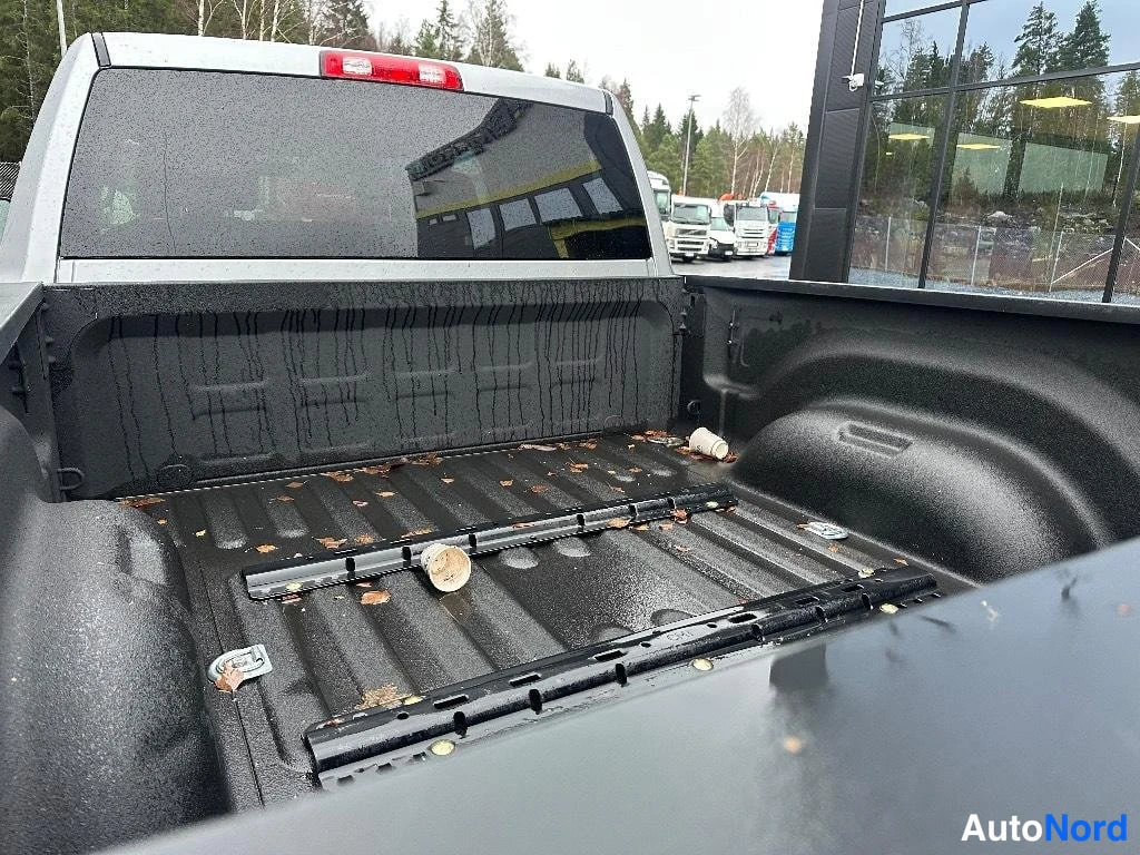 Yksityiskohta: Lavan sisäpinta harmaassa lava-autossa, musta muovipinnoite ja kaksi mustaa kiinnityskiskoa, lehtiä ja roskia lavassa.