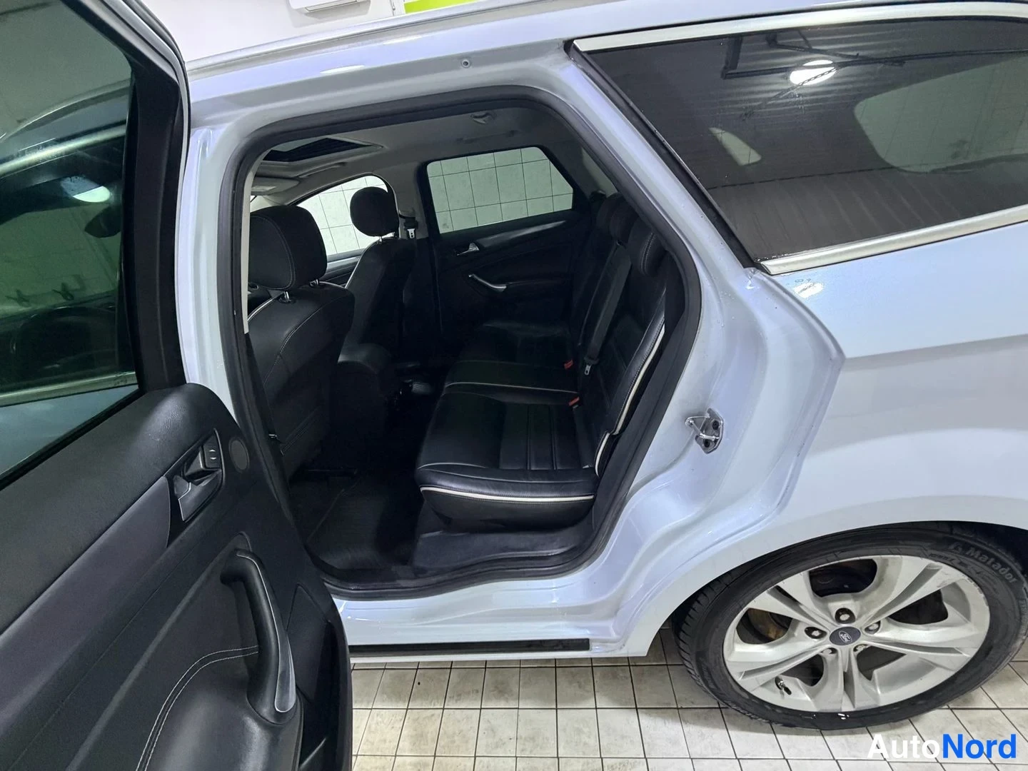 Muu: Takaoven aukaisema auto: näkyvät mustat takapenkin istuimet ja valkohelmattu nahkapinta, hopeanvärinen alumiinivanne, sadeveden poistokouru takaluukussa.