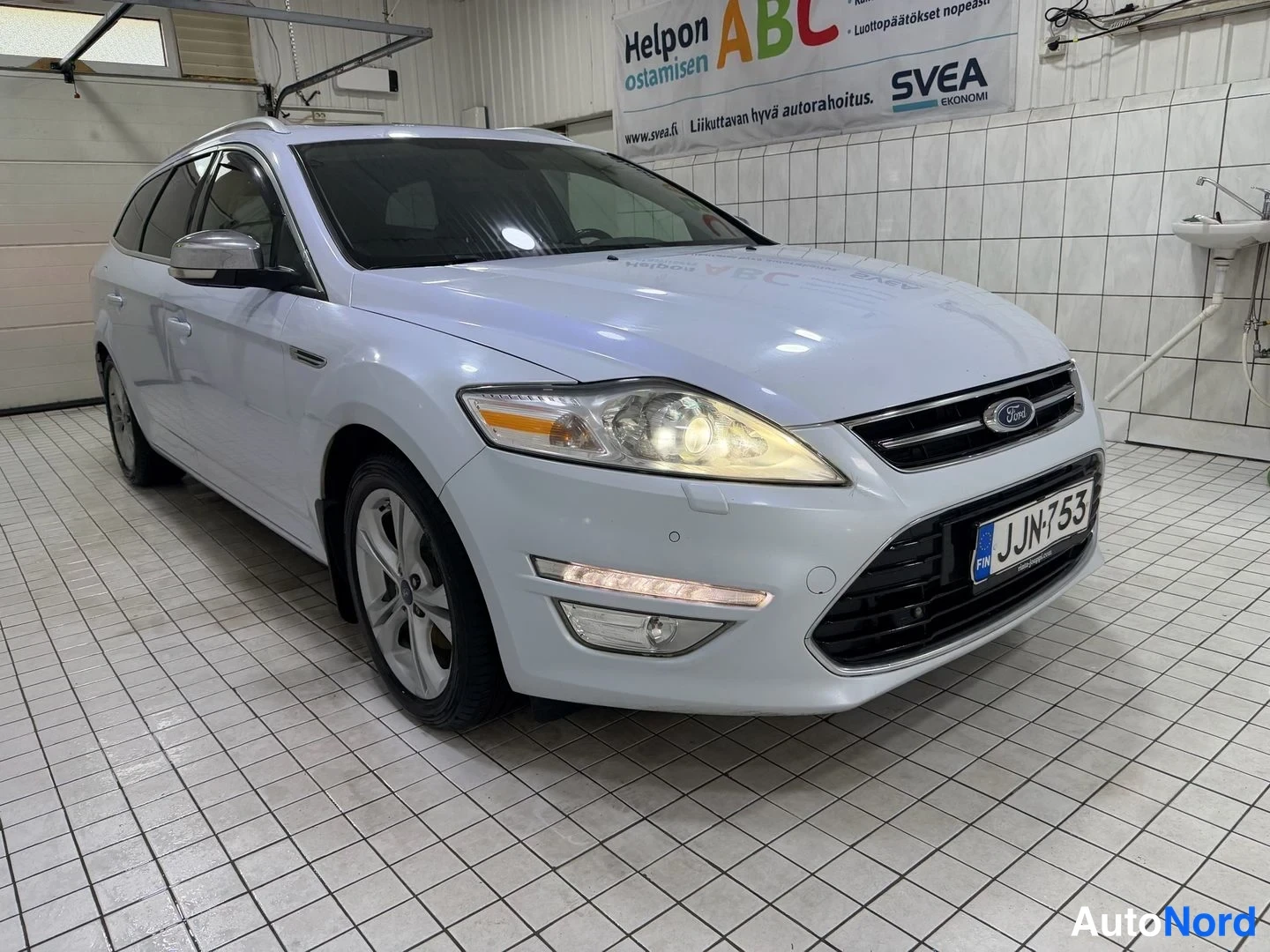 Etuviisto, nokka oikealle: valkoinen Ford Mondeo Farmari etukulmasta oikealta kuvattuna, rekisterikilpi, kattokaiteet näkyvät; auto parkissa laattalattialla.