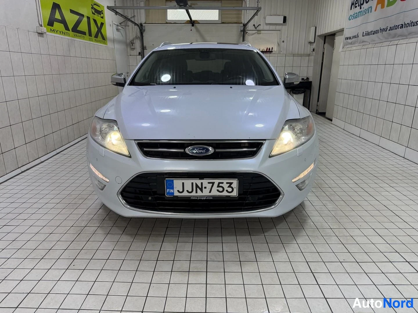 Edestä: Ford Monoco... valkoinen farmari edestäpäin, rekisteritunnus, ajovalot päällä.