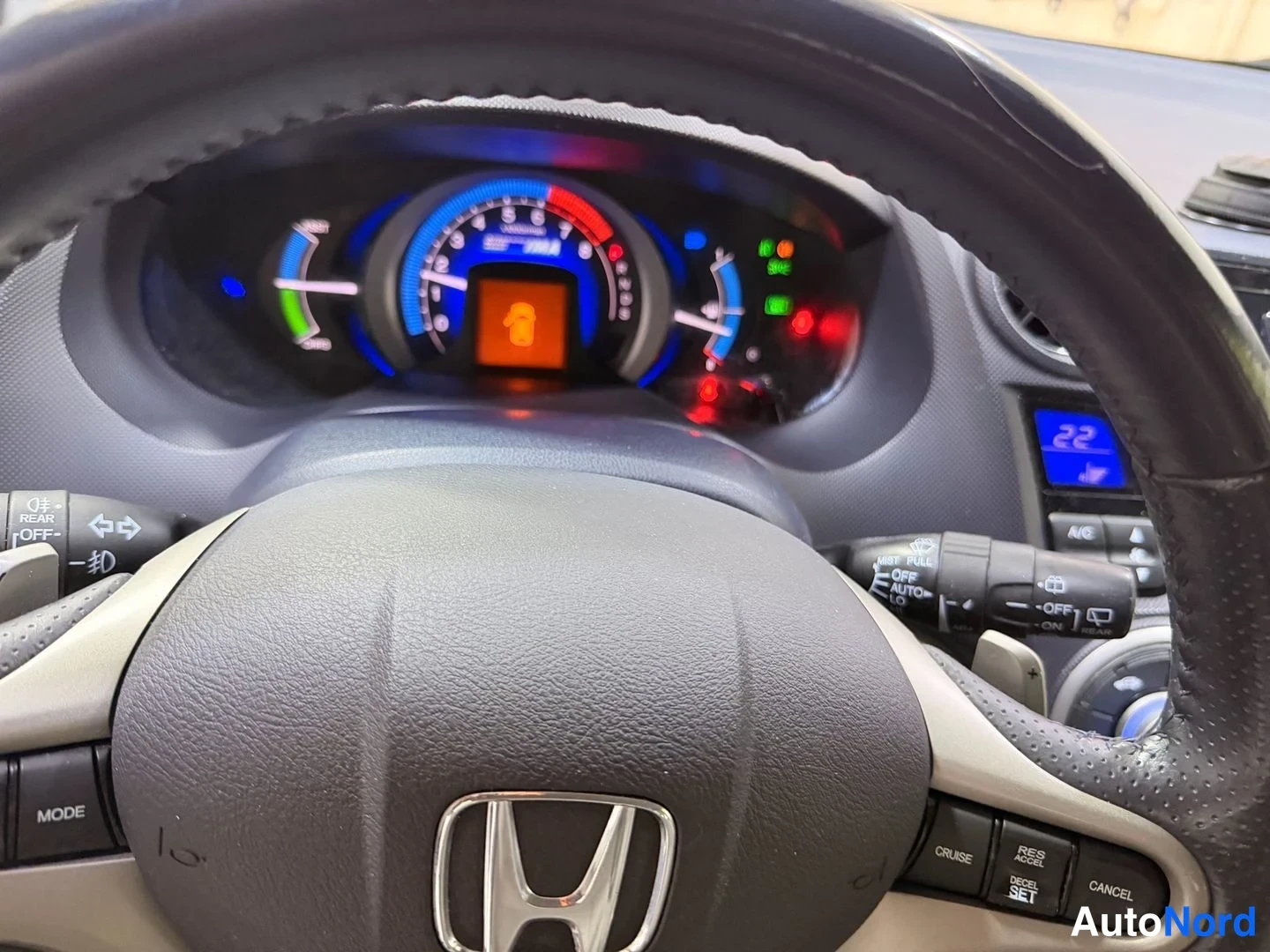 Muu: Honda-merkillä varustettu ohjauspyörä ja mittaristo, jossa näkyvät nopeusmittari, ajovalot ja varoitukset; infonäytössä lukee 22.