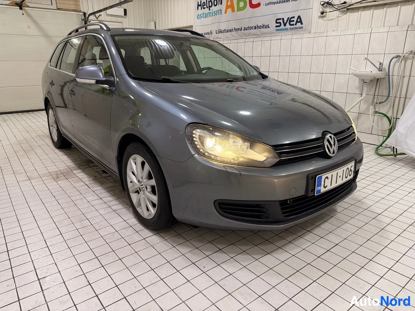 Etuviisto, nokka oikealle: Farmarimallinen henkilöauto etukulmasta oikealta kuvattuna; etusäleikkö, Volkswagen-merkki, toisessa etulyhdyssä hehkuva valo ja etupuskurissa rekisterit