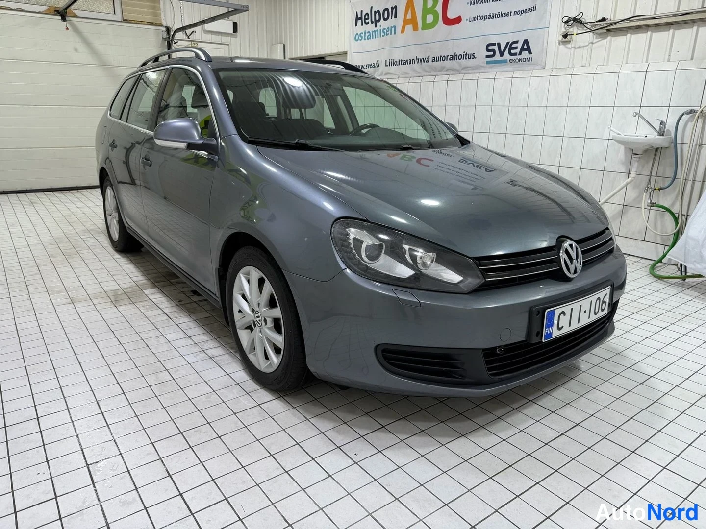 Etuviisto, nokka oikealle: Auton keula ja kylki kolmeneltasojen kulmasta; harmaa farmari, etusäleikkö, Volkswagen-merkki, rekisterikilpi ja kattokaiteet.