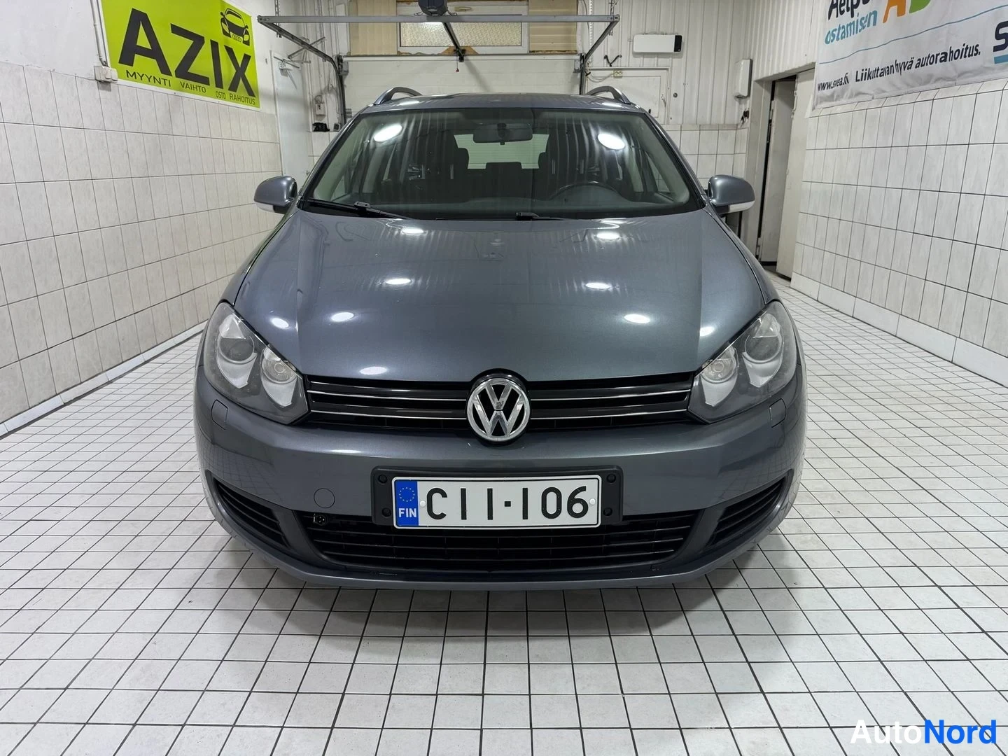 Edestä: Auton keula suoraan edestä; harmaa Volkswagen, etusäleikkö, merkki, takavalojen ja kaukovalojen välissä rekisterikilpi, kattokaiteet katolla.