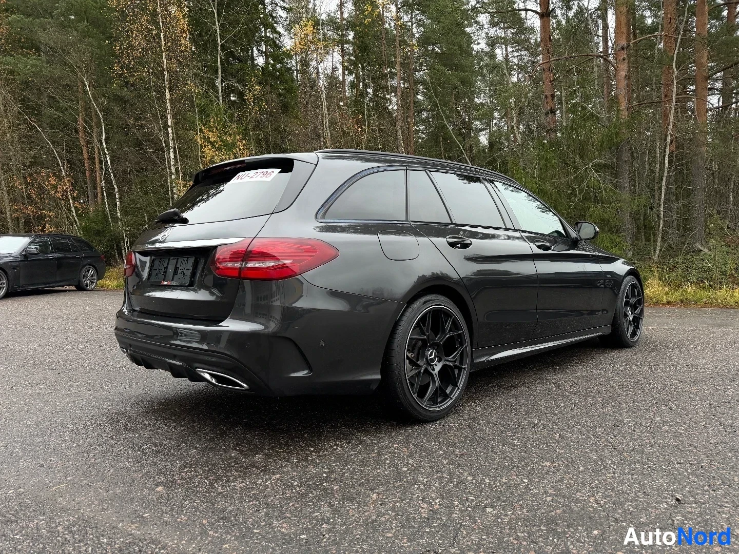 Takaviisto: Auton takapää ja vasen sivu kolmannesnäkymässä: tummanharmaa farmariauto tummilla vanteilla asfalttipihalla, metsä taustalla.