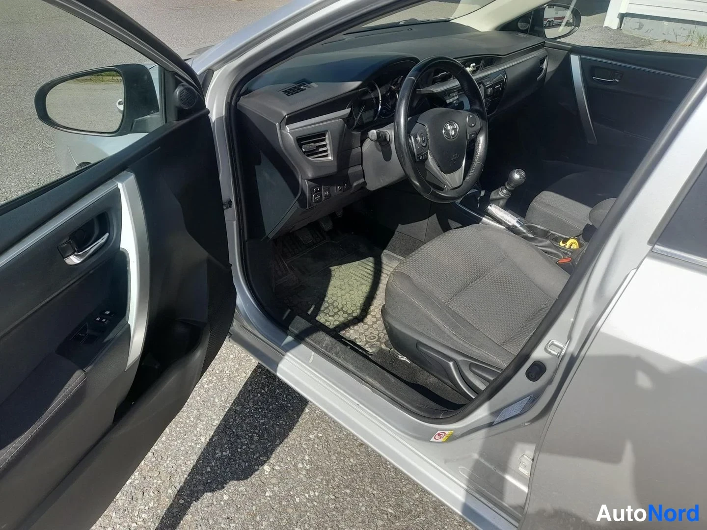 Sivulta: Toyota Corollan kuljettajan ovi auki ja ohjaamo näkyvät: ratissa Toyota-merkki, käsivaihtoja ja tummat penkkiistuimet.