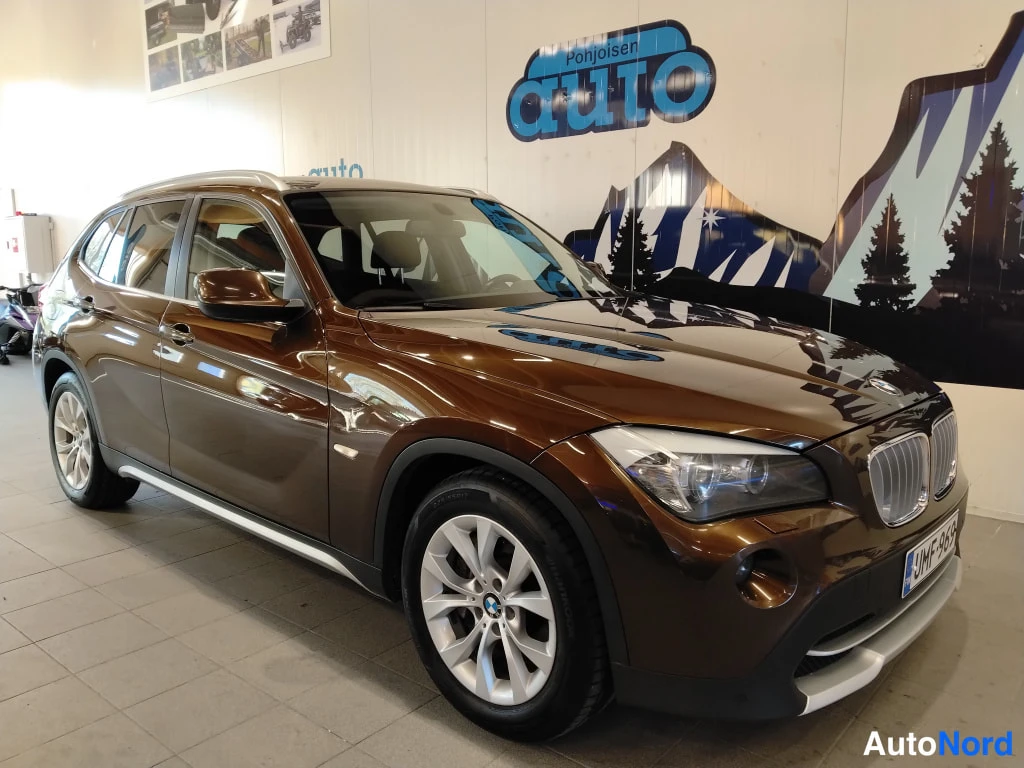 Etuviisto, nokka oikealle: BMW X1 -automobiilin etukulman punos kuvattuna sisätiloissa; auto on ruskeakyllä, eikä tankia näy.