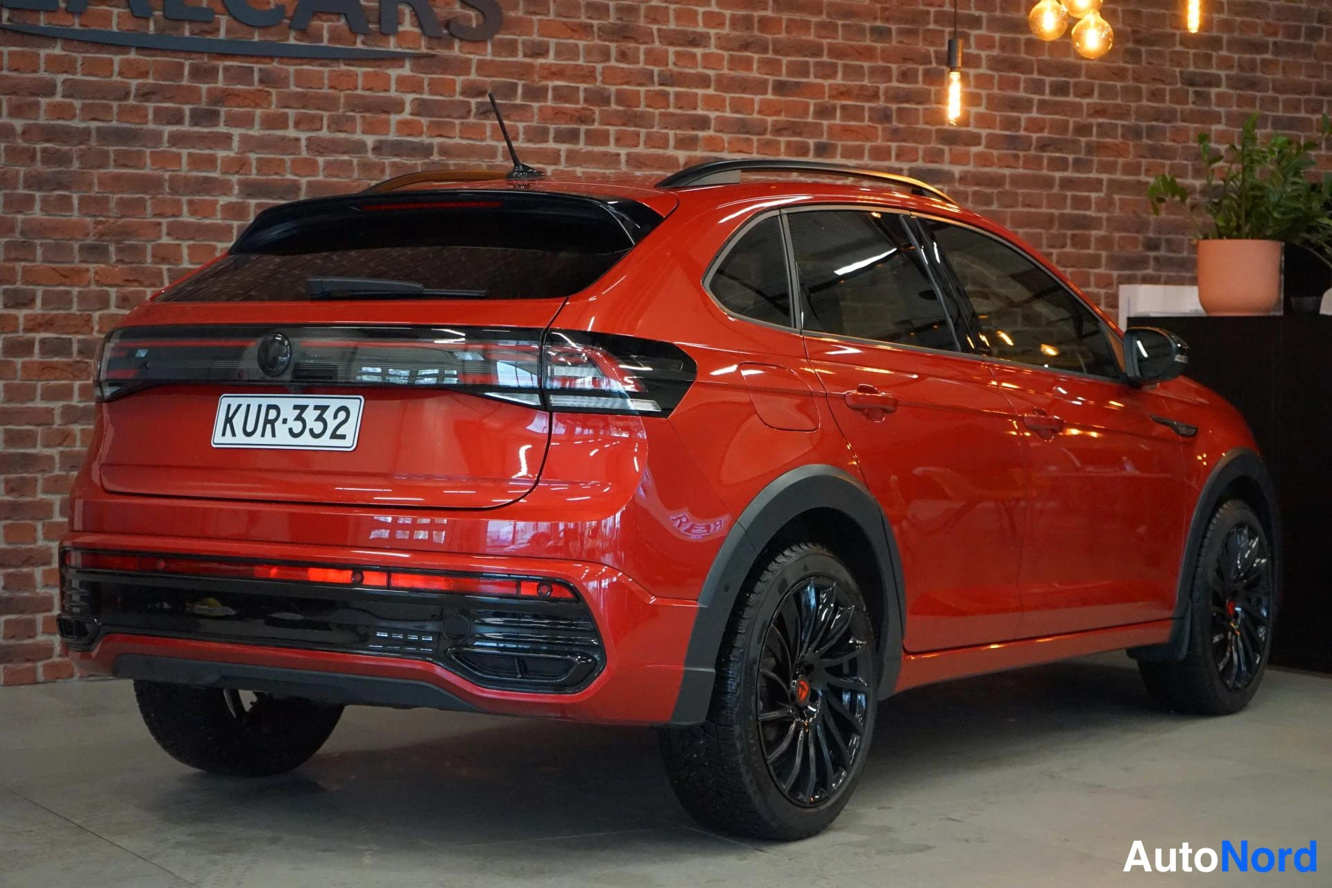 Takaviisto: Punaisen Volkswagen Taigon takaosa kylki näkyvissä, rekisterikilpi, tummat takaovat ja mustat vanteet tiiliseinää vasten.