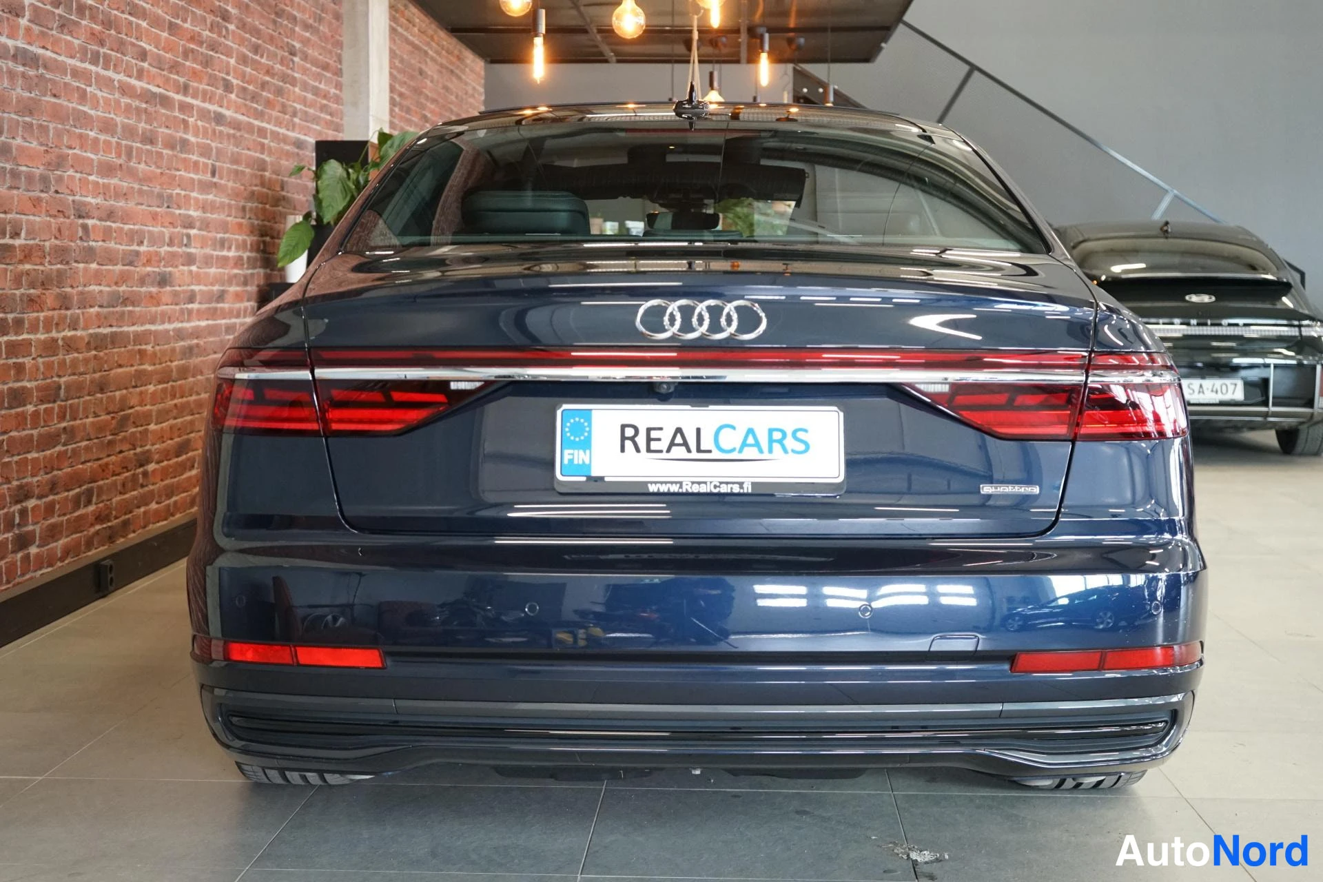 Takaa: Audi A8in takaosa suoraan takaa: tumma kori, punaiset takavalot vaakakuviona, neljän renkaan merkki ja RealCars-kyltti takalokin päällä.