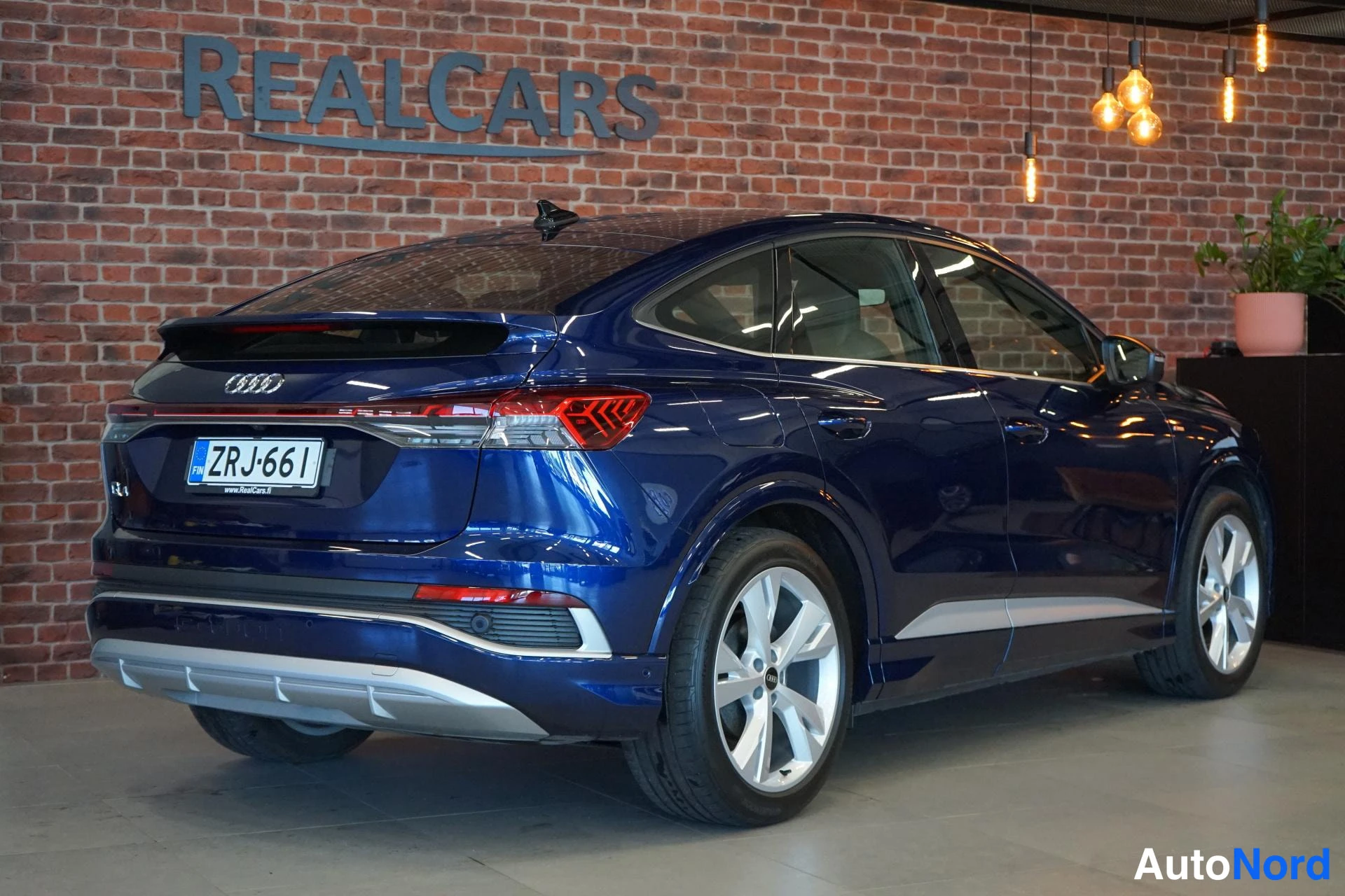 Takaviisto: Tummansinisen Audi Q4 e-tron Sportbackin takaosa kolmesta neljänneskulmasta; takavalojen kauttakulkeva vaakavalotanko ja rekisterikilpi näkyvät tiiliseinää vast