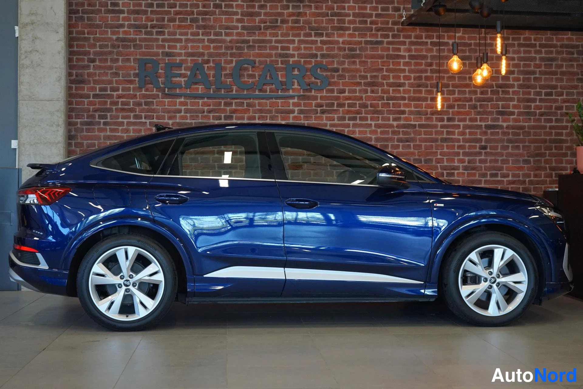 Sivulta: Tummansinisen Audi Q4 e-tron Sportbackin sivuprofiili sisätiloissa; avaimenreiätön avaus ja hopeiset ala listat näkyvät ovissa.