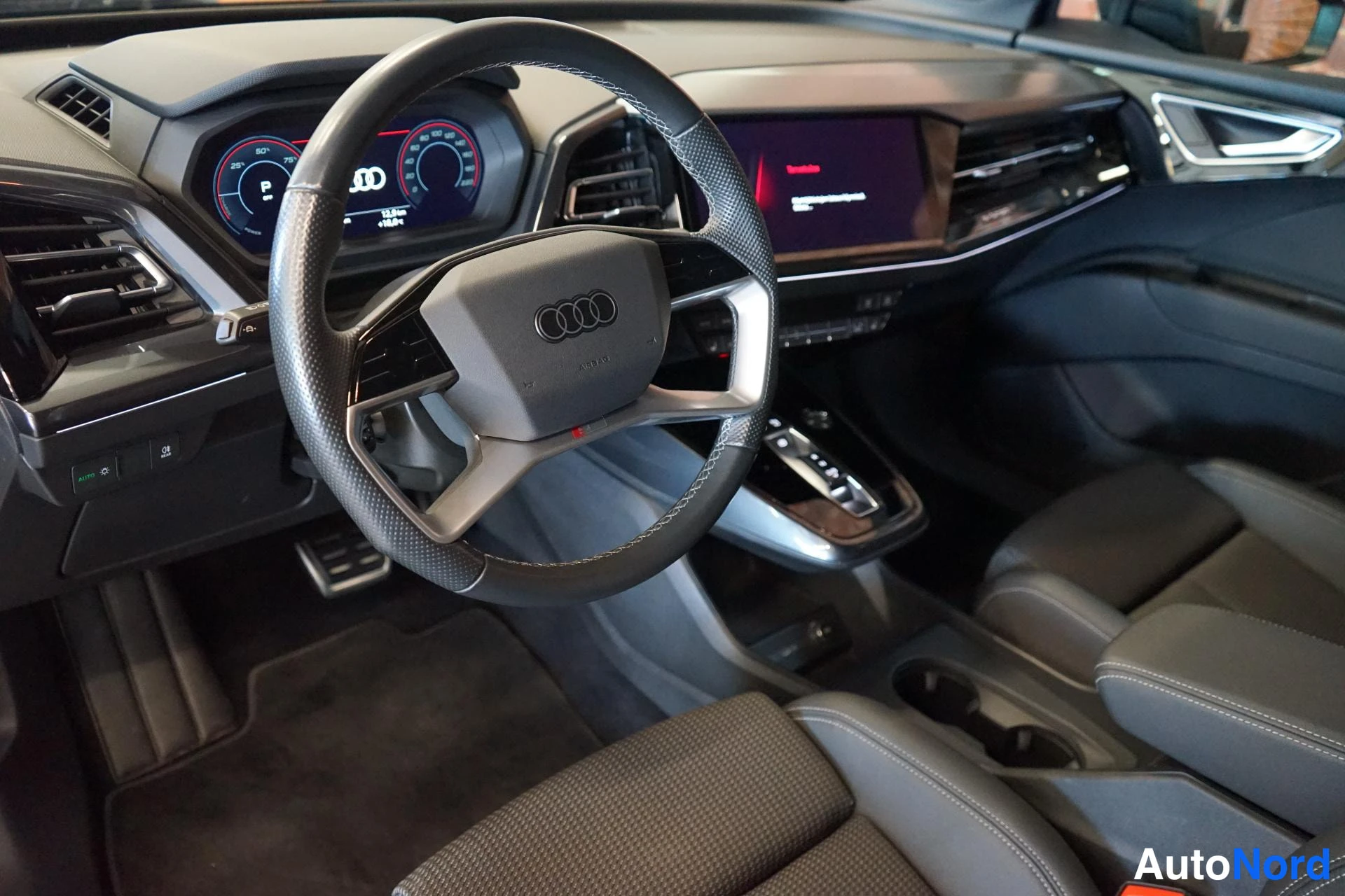 Muu: Auton sisällä ohjauspyörä Audi-tunnuksella, digitaalinen mittaristo, keskikonsolen kosketusnäyttö ja tummat etuistuimet.