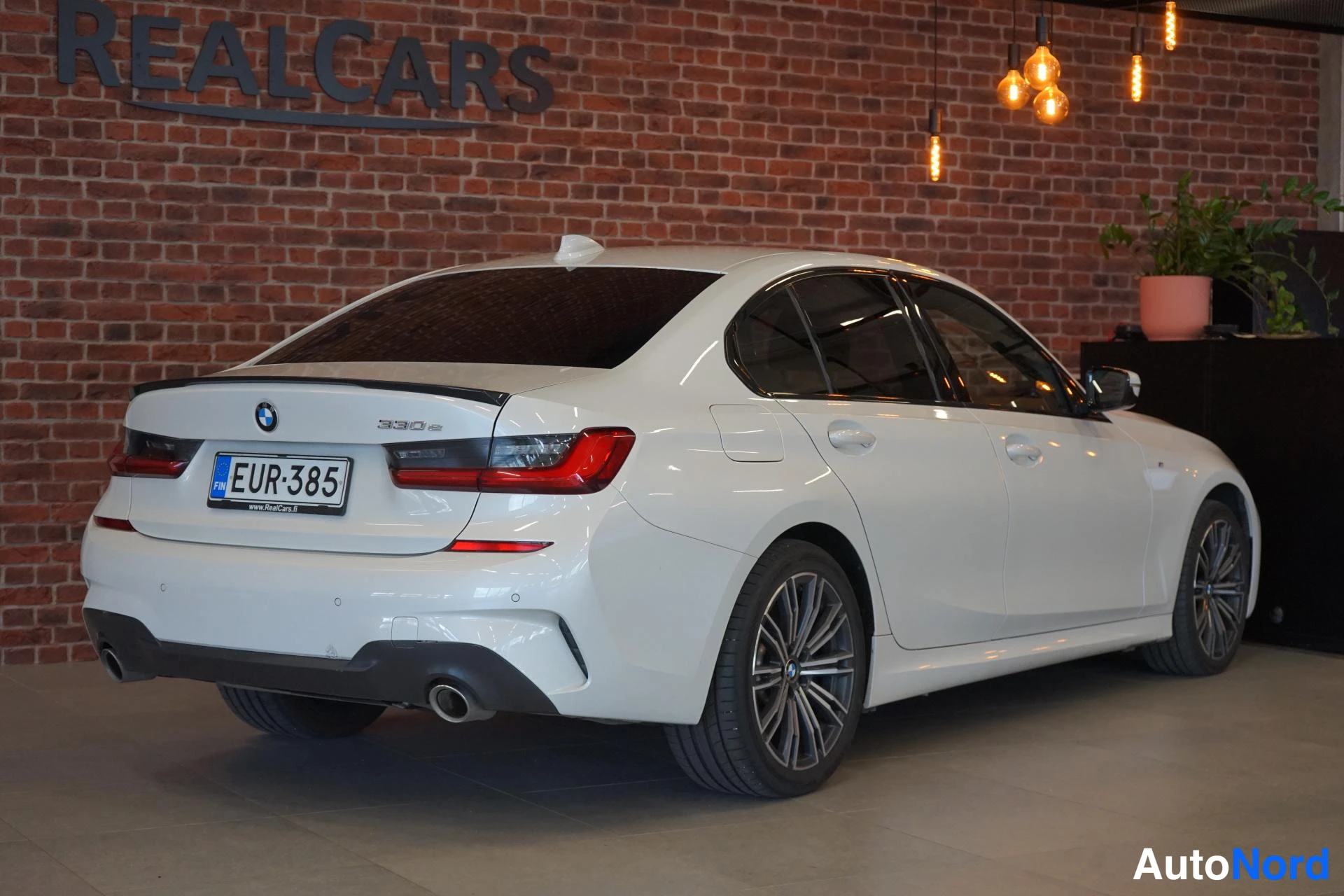 Takaviisto: Valkoinen BMW-sedan takaosa kolmeneljäsosakulmasta, takavalojen rekisterikilpi ja kaksoispakoputki näkyvät, takana tummennetut ikkunat.