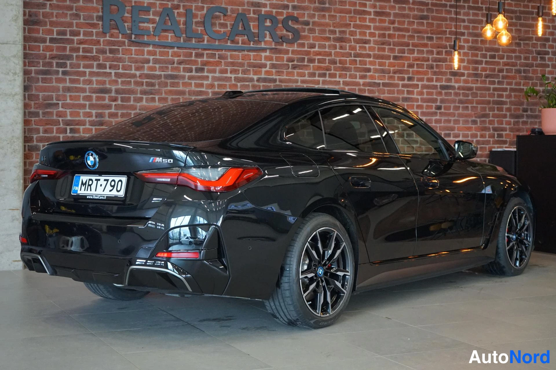 Takaviisto: Mustan BMW:n takaosa ja kylki kolmestavartsesta sisätiloissa tiiliseinä taustalla, rekisteritunnus.