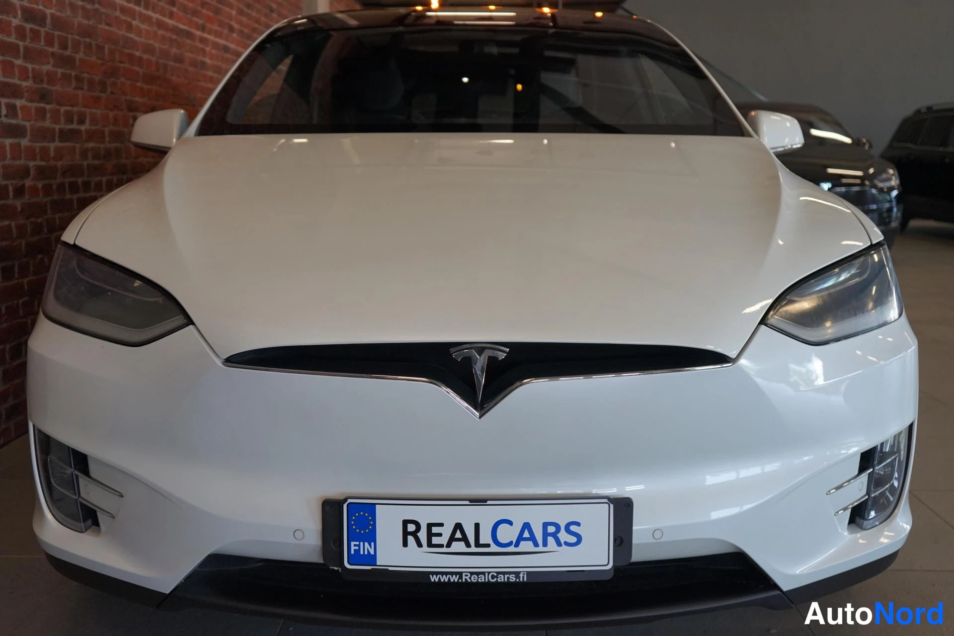 Edestä: Valkoisen Tesla Model X:n keulan lähikuva, T-merkki ja TESLA-logo konepellin nokassa näkyvät; tuntematon malli.
