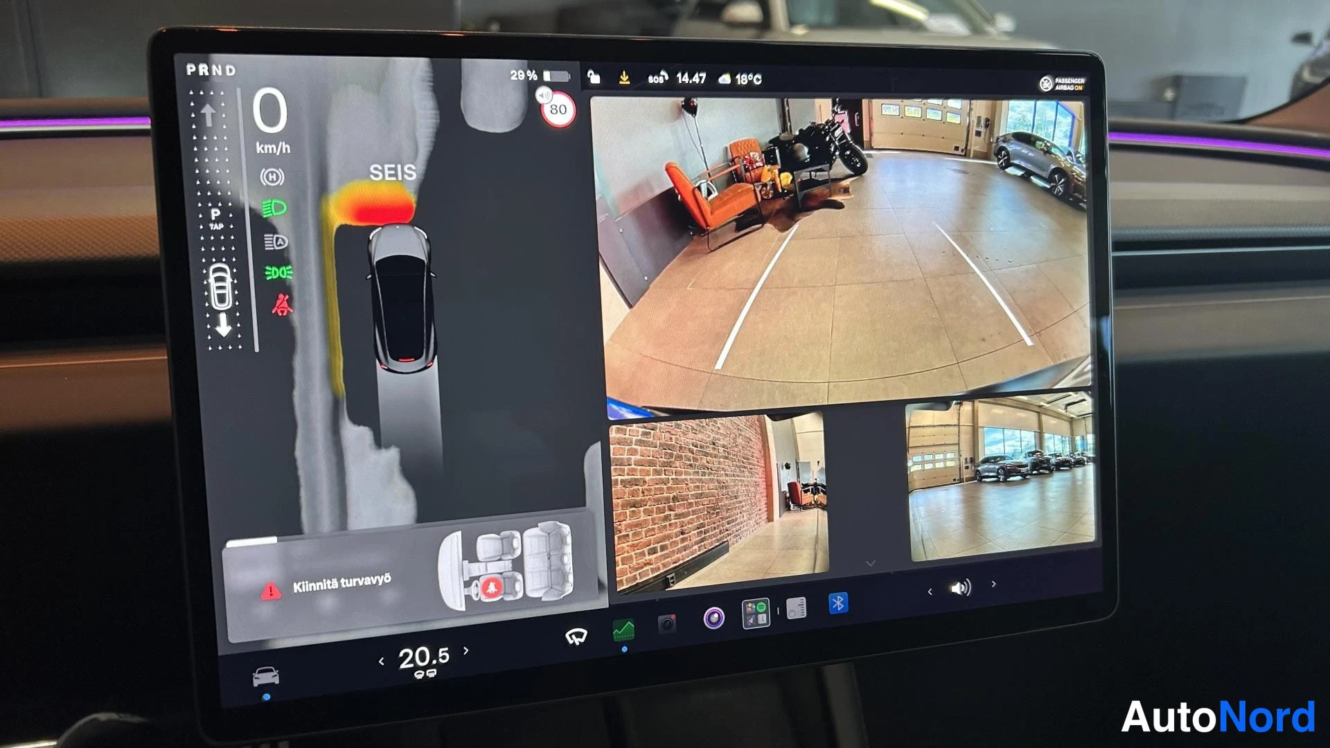 Muu: Tesla Model 3:n keskinäyttö, jossa näkyy pysäköintikameranäkymä, Ford-korjaamon seinäkilpi ja varoitus 'Kiinnitä turvavyö'.