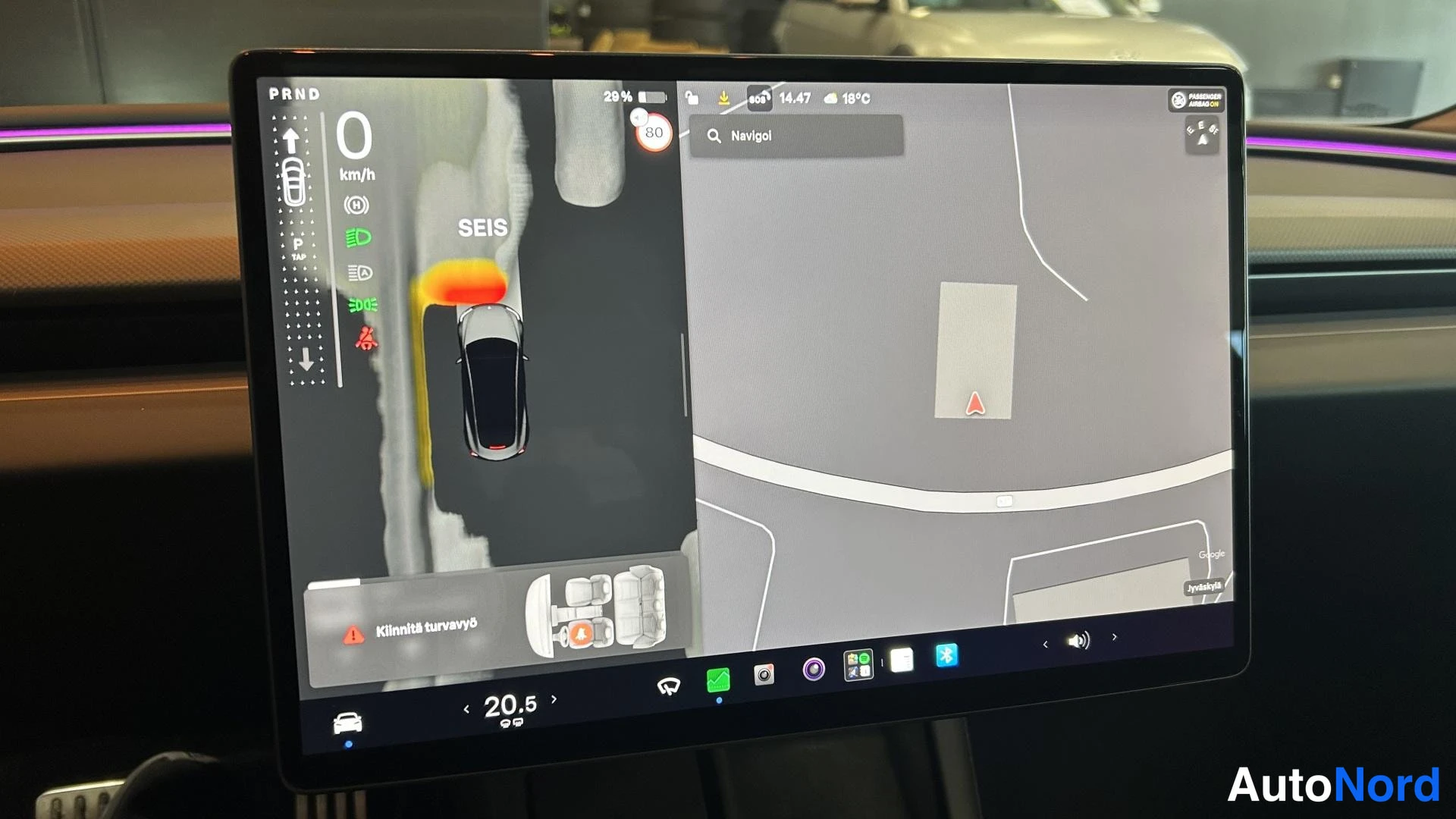 Muu: Tesla Model 3:n keskinäyttö karttanavigointiohjelmalla ja pysäköintitilanteen visualisoinnilla, varoitus 'Kiinnitä turvavyö' alareunassa.