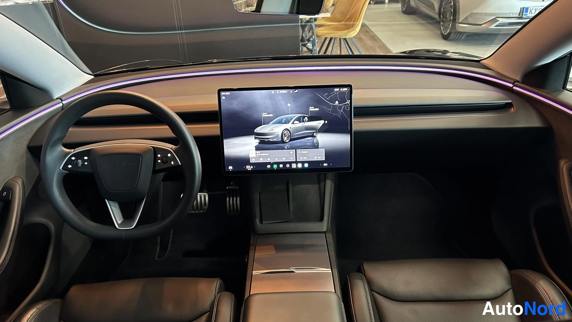 Muu: Tesla Model 3:n ohjaamo edestä: keskellä suuri keskinäyttö, vasemmalla ohjauspyörä, tummat verhoilut ja violettia valoa antava tunnelmavalo kojelaudan yläreunas