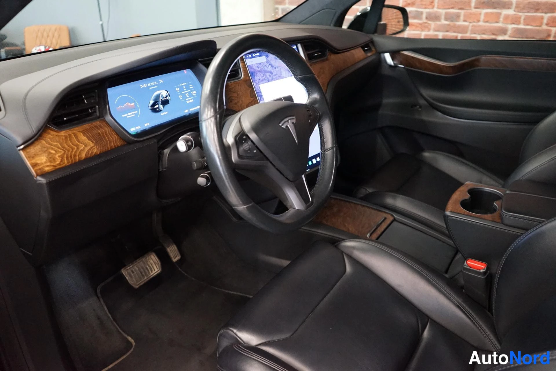 Edestä: Tesla Model X:n ohjaamo: puukoristeinen kojelauta, keskikonsolen iso kosketusnäyttö ja mustat nahkaistuimet.