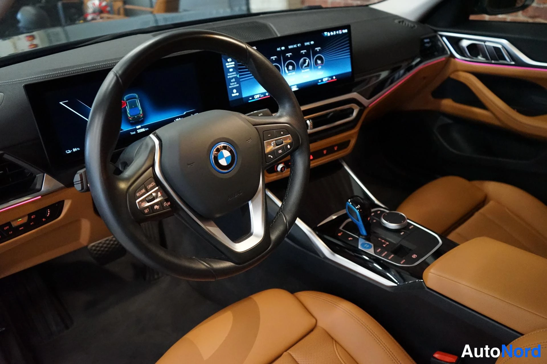 Muu: BMW:n ohjaamon näkymä: ratissa BMW-logo, pitkä näyttökojelaudan ja infotainment-näyttö, ruskeat nahkaistuimet ja keskikonsoli.