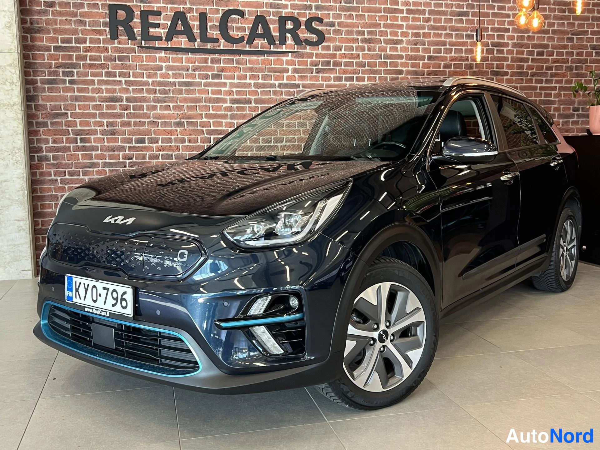 Etuviisto, nokka vasemmalle: Musta Kia Niro Electric -henkilöauto etuviistossa showroomissa, tiiliseinällä REALCARS-teksti.