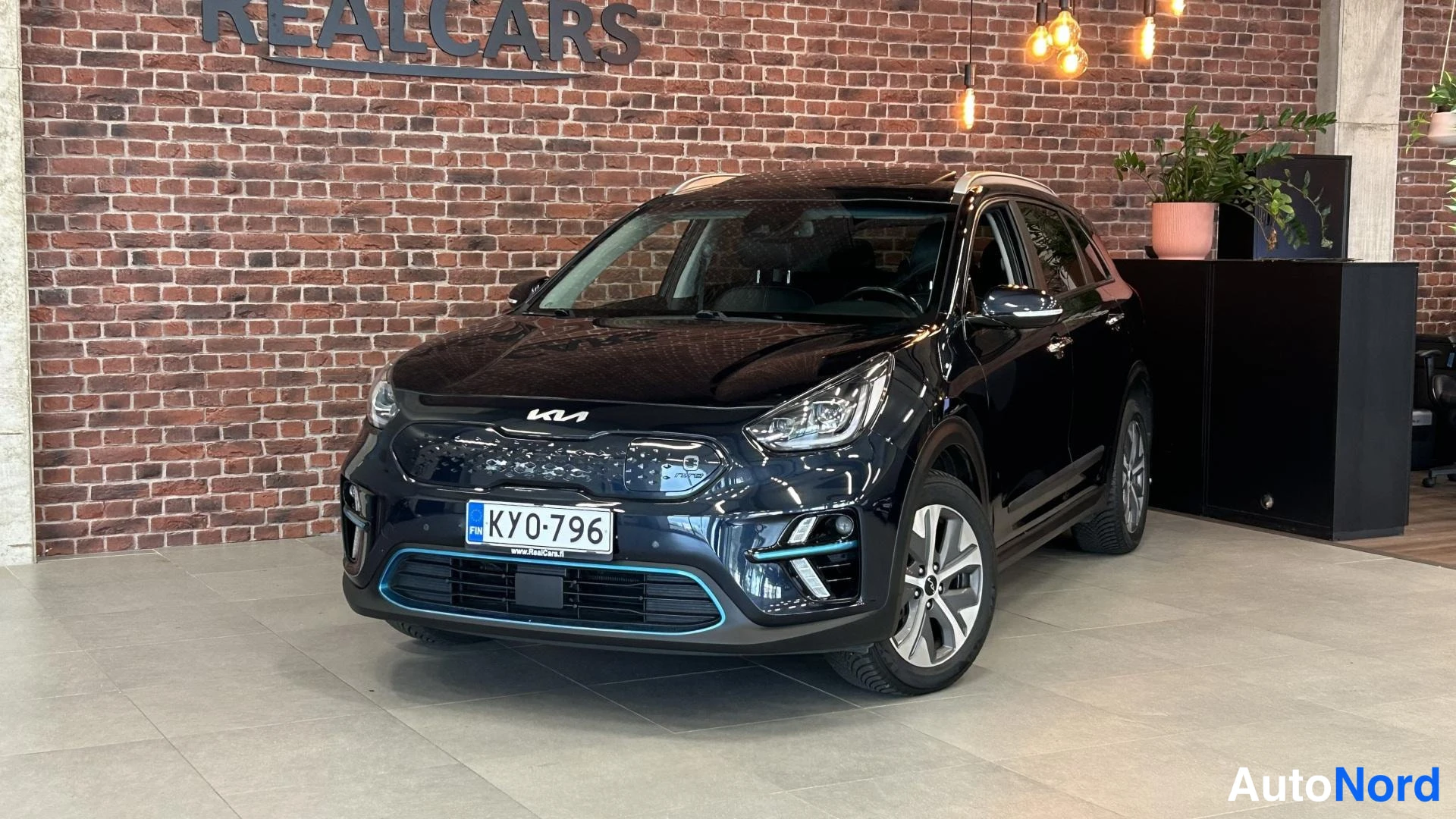 Etuviisto, nokka vasemmalle: Musta Kia Niro Electric -sähköauto myyntihallissa, edessä kolmannesvasemmalla profiilissa.