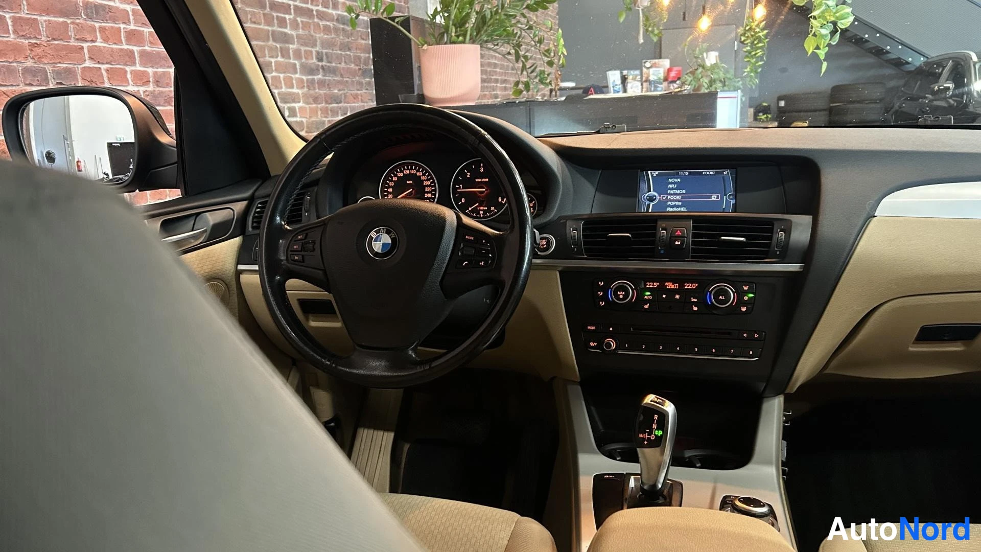 Muu: Autojen ohjaamo sivulta: BMW-ohjauspyörä, mittaristo, keskikonsoli ja automaattivaihteiston valitsin, vaaleat istuimet.