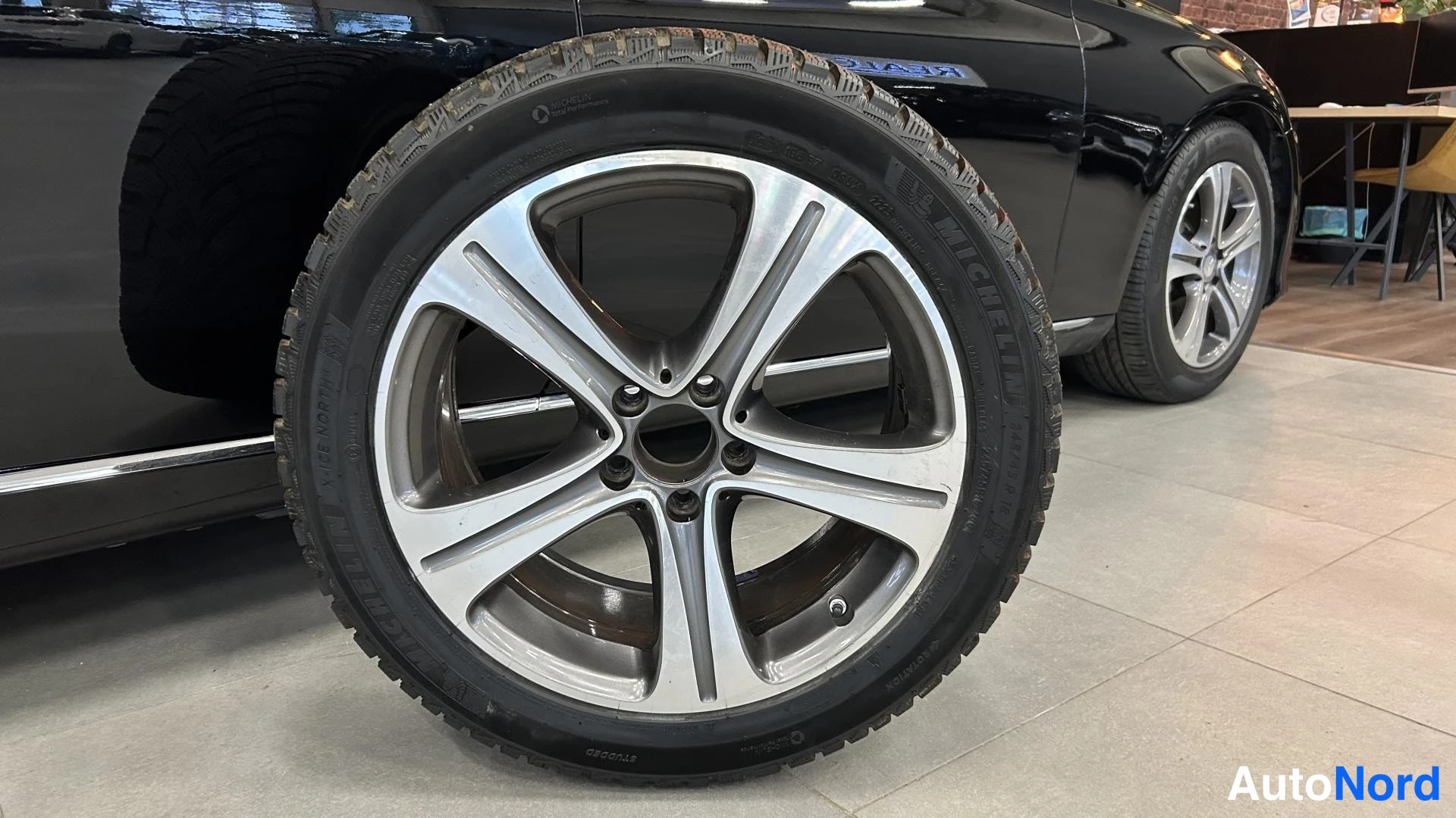 Sivulta: Musta henkilöauto ja irrotettu alumiinvanne kokotpayrellä, rengasmerkintä Michelin, myyntitilan lattialla.