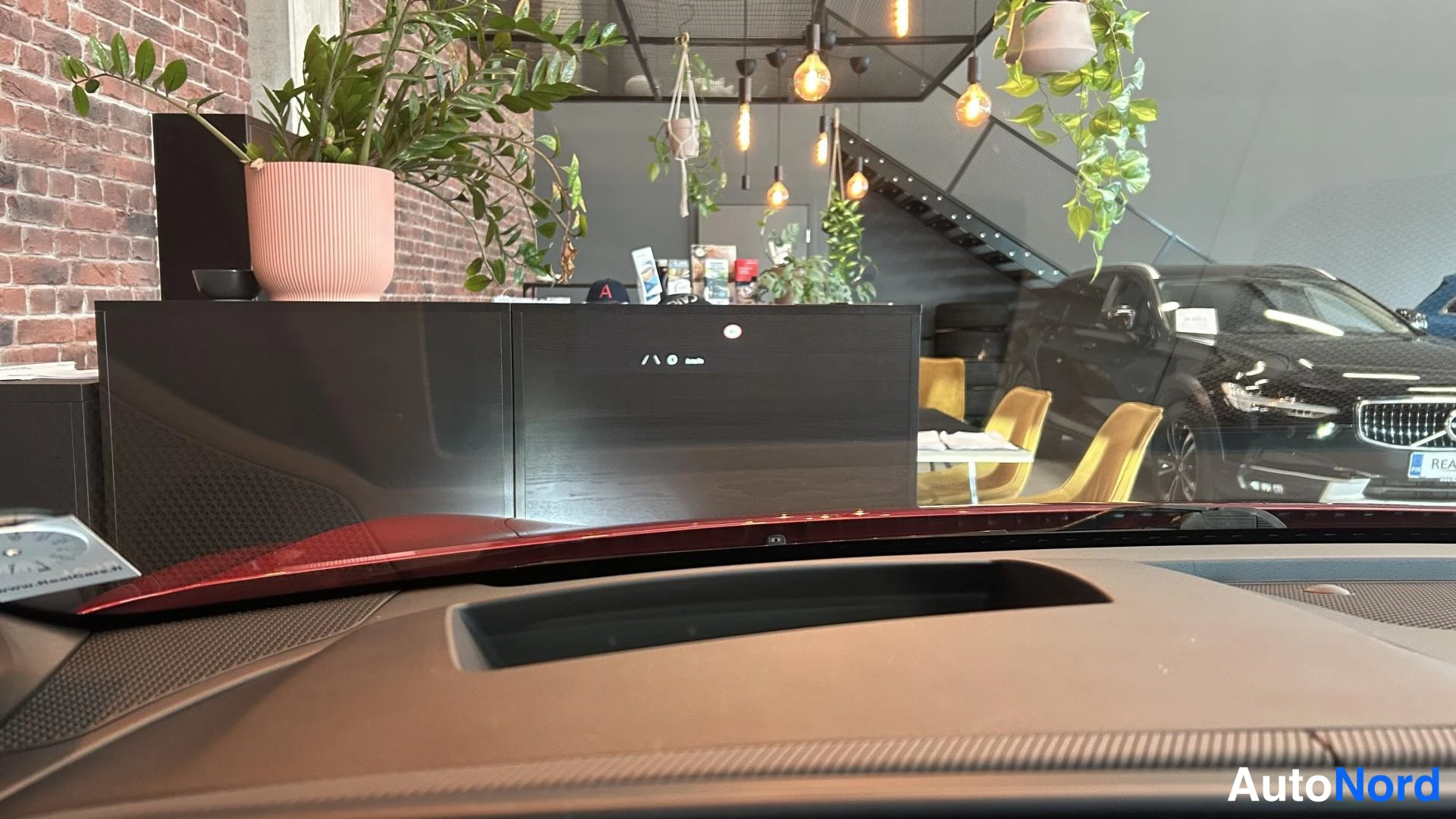 Muu: Näkymä auton kojelaudan tuulilasipuolelta: tumma dashboard, takanäkyvissä myymälähuone, istutuskorussa kasvit ja kelttuolit, joiden takana musta henkilöauto.
