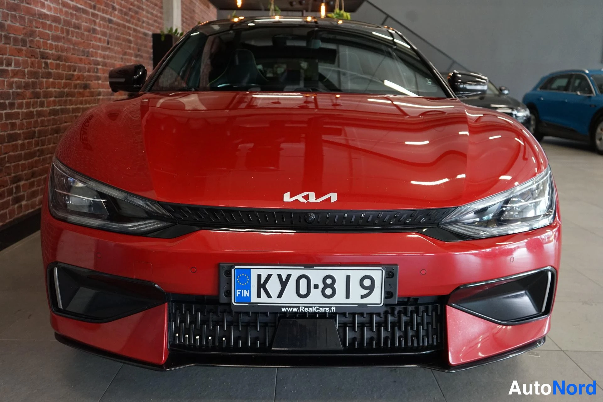 Edestä: Punaisen Kia EV6 -auton etunäköympyrä: Kia-merkki konepellissä, rekisterikilpi KY0-819 ja www.RealCars.fi -teksti.