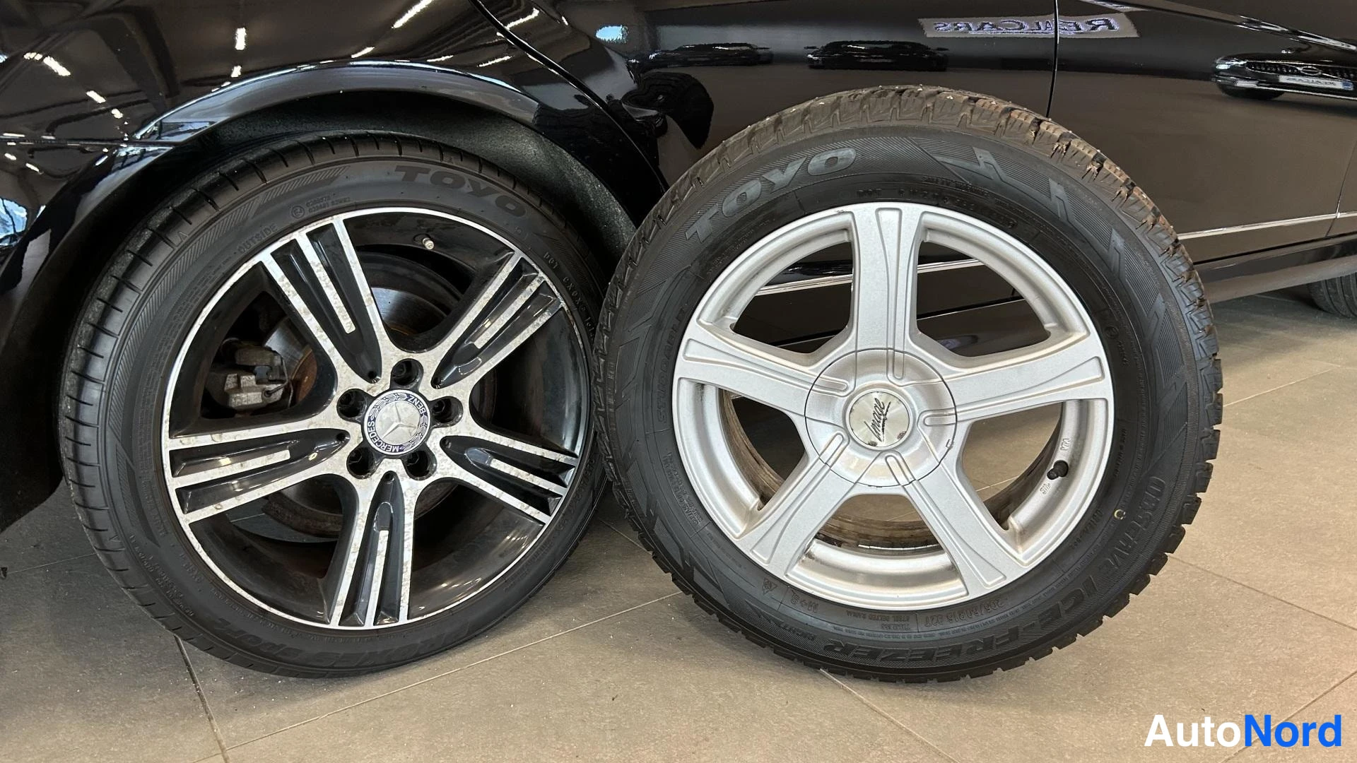 Yksityiskohta: Kaksi irrotettua rengasta vanteilla: vasemmalla tumma Mercedes-merkitty monipuikko vanne, oikealla hopeinen viisipuikkovanne TOYO-renkaalla, lattialla autotalli