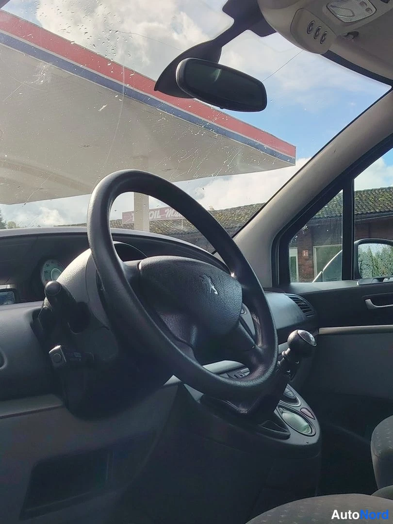 Muu: Interior view of the dashboard with steering wheel and manual gear lever, photographed through a rain-spotted windshield showing a gas station canopy.