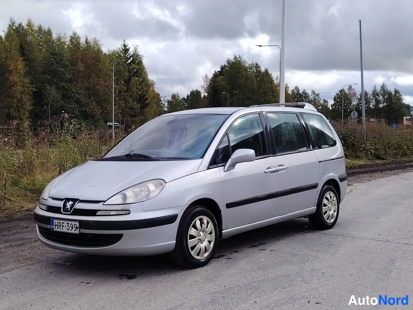Etuviisto, nokka vasemmalle: Hopeanvärinen tila-auto kuvattu etukolmisesta vasemmalta; rekisterikilpi, kattotelineet ja hopea maali.