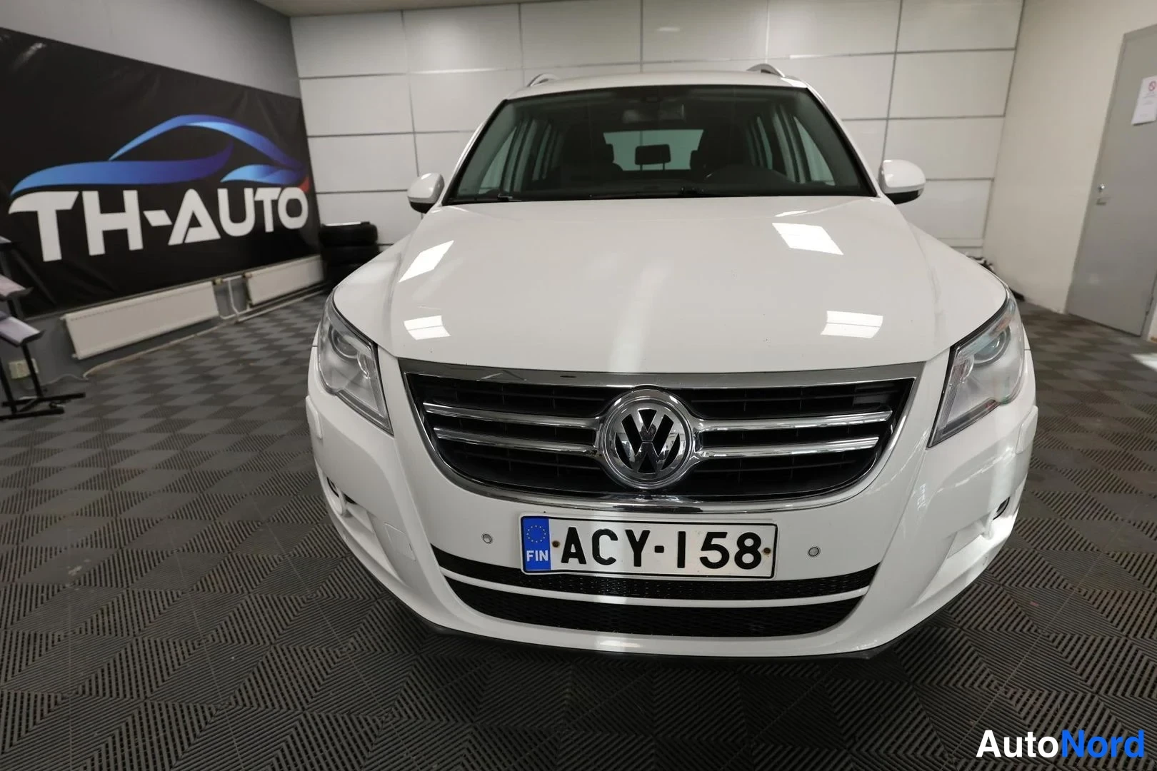 Edestä: Valkoisen Volkswagen Tiguan etunäkymä sisätiloissa, rekisterikilpi ja VW-merkki näkyvissä.