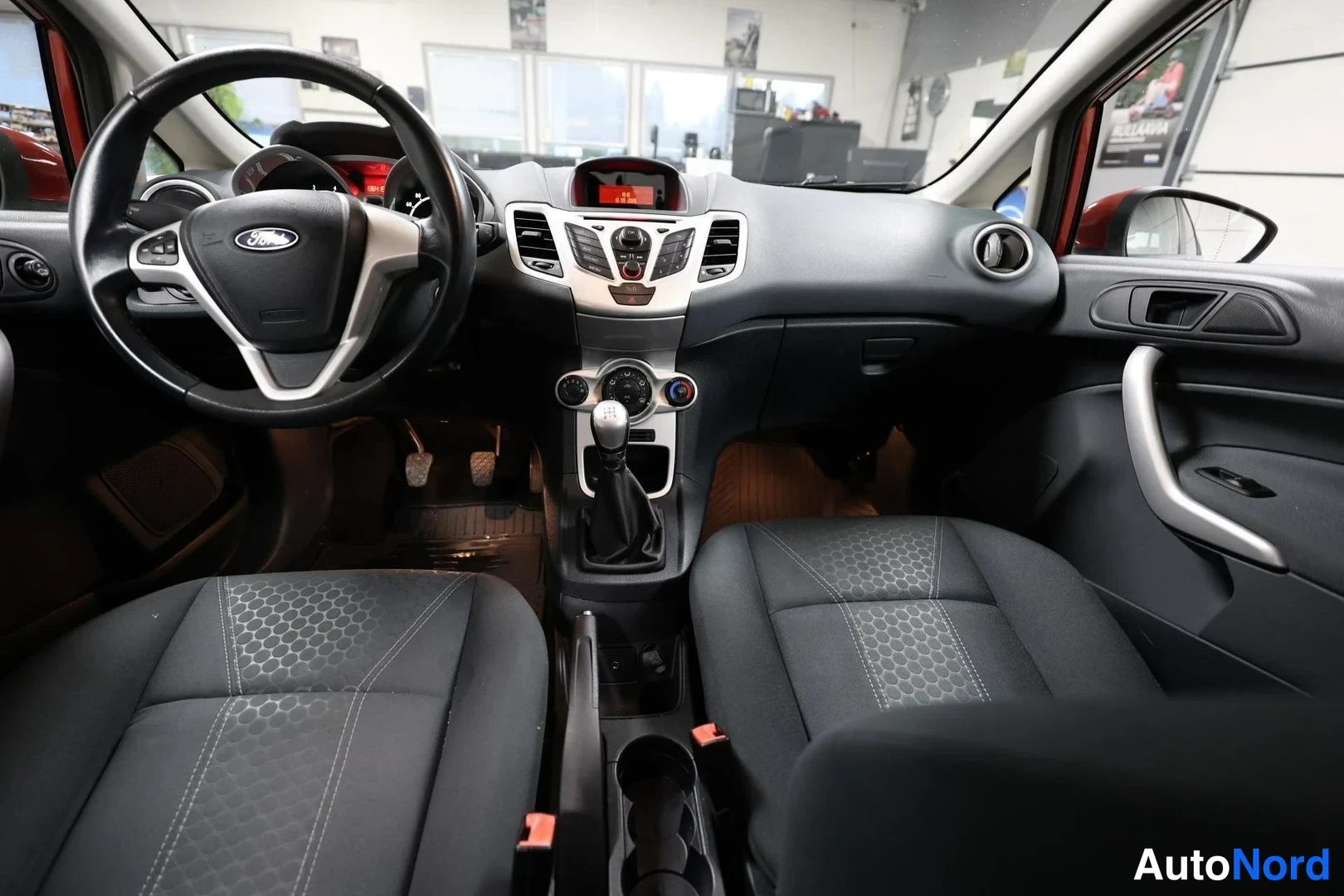 Muu: Ford Fiesta -ohjaamo etukädestä: ohjauspyörä, käsiradio, manuaalivaihteiskeppi ja kaksi etuistuinta tummalla kankaalla.