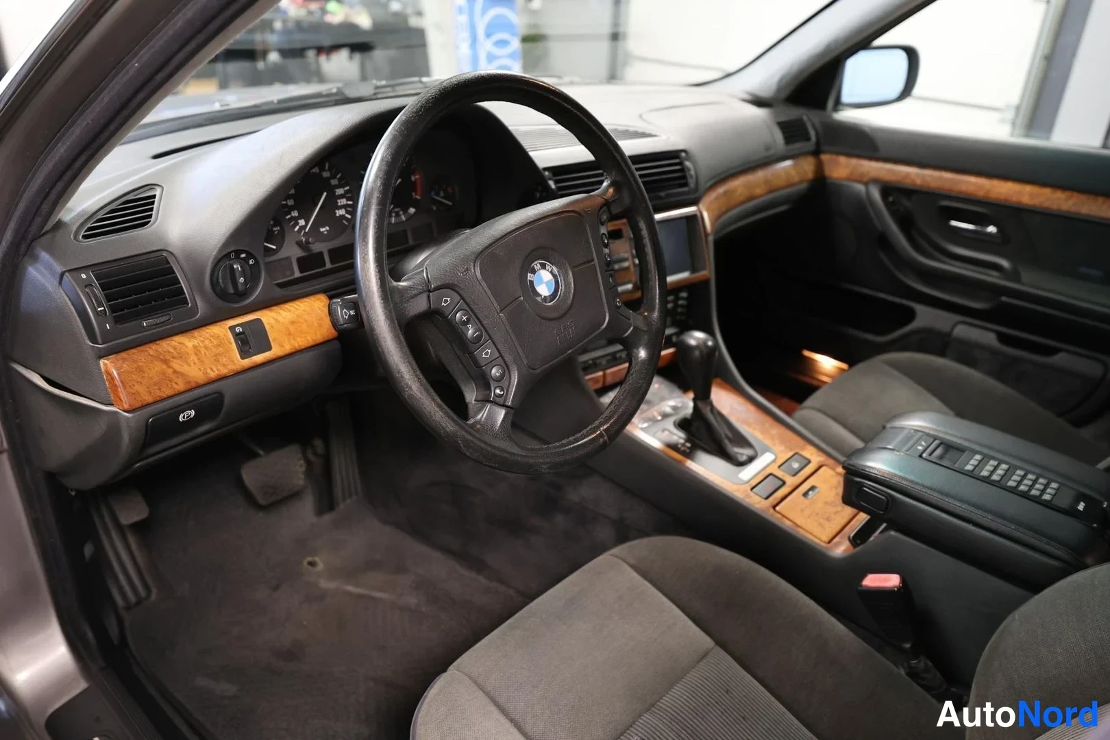 Muu: Auton sisätilat: musta ohjauspyörä BMW-merkillä, instrumentit, puukoristeinen kojelauta ja vaihtajanäkymä kuljettajan puolelta.