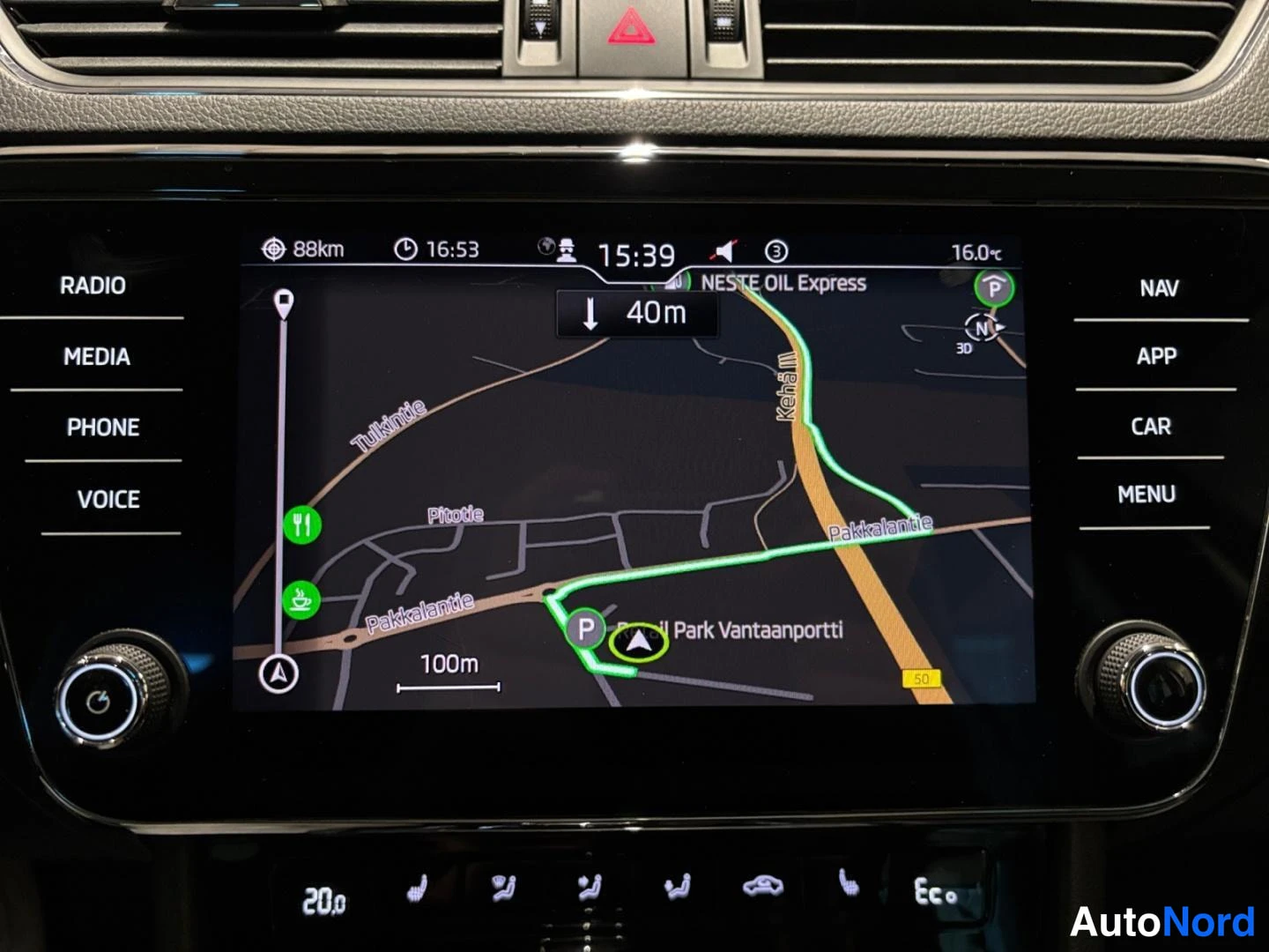 Muu: Auton kojelaudan infotainment-näyttö näyttää navigointikarttaa reittiohjeineen; ympärillä RADIO-, MEDIA-, PHONE-, VOICE-, NAV-, APP-, CAR- ja MENU-painikkeet.