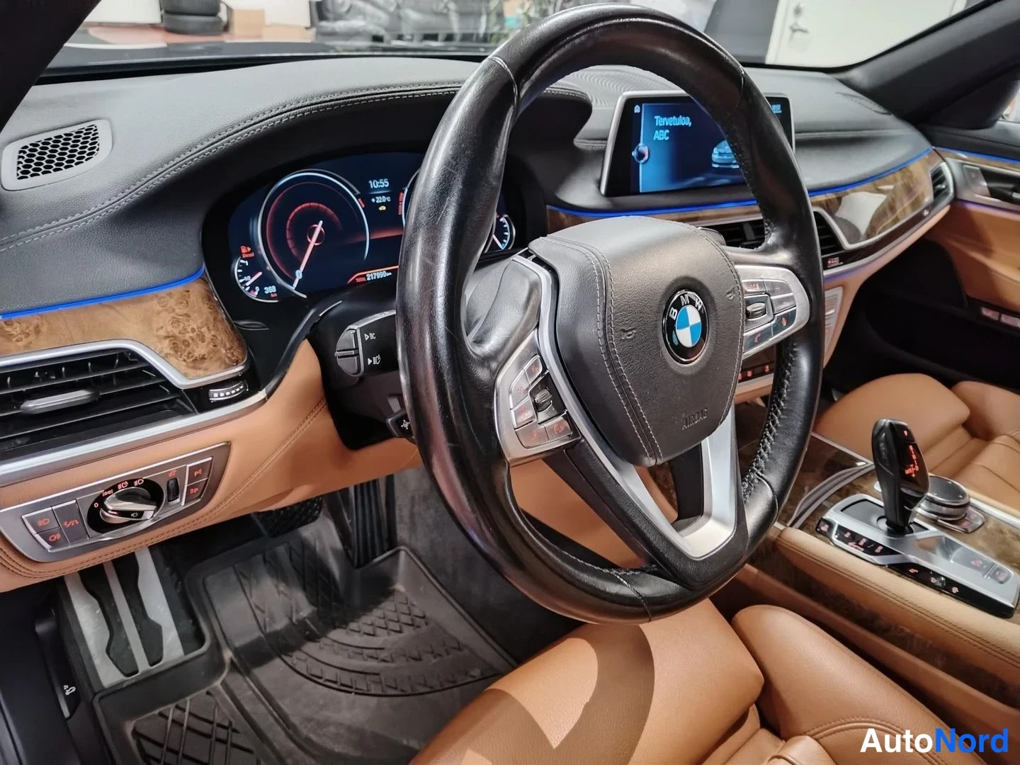 Muu: Sivukulma auton ohjaamosta: BMW-ohjauspyörä lähikuvassa, digitaalinen mittaristo, vasemmassa reunassa ilmastointisäätimet ja oviverhoilu ruskeassa nahassa.