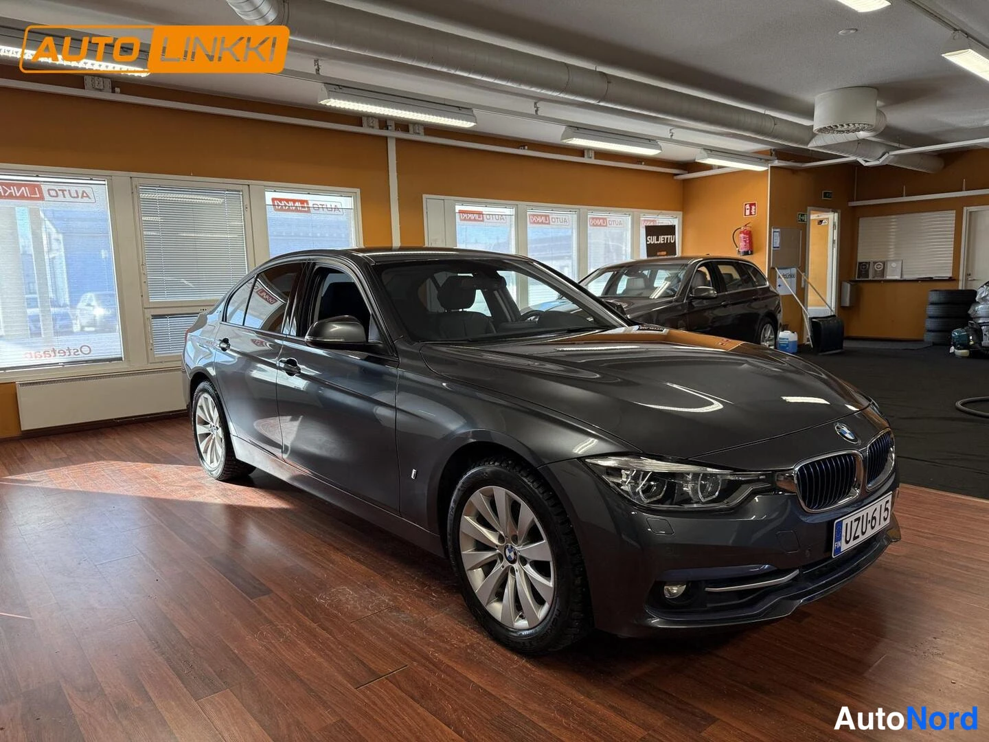 Etuviisto, nokka oikealle: Tummanharmaa BMW-sedan etukulmasta sisätiloissa, rekisterikilpi.