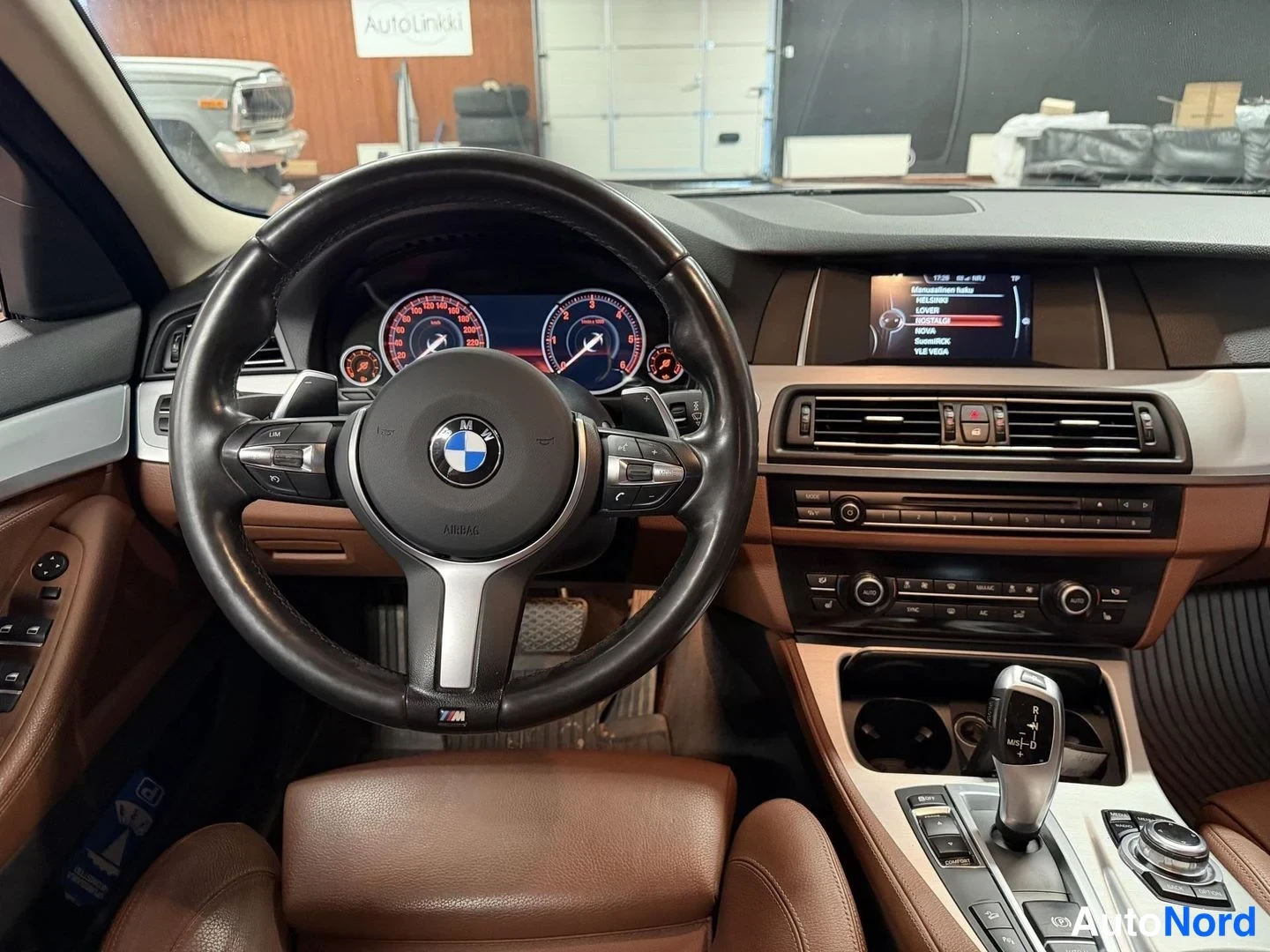 Muu: Ohjaamo, BMW-merkkinen M-ohjauspyörä ja ruskeat nahkaistuimet; vaihteenvalitsin ja iDrive-ohjain keskikonsolissa, näytöllä valikko.