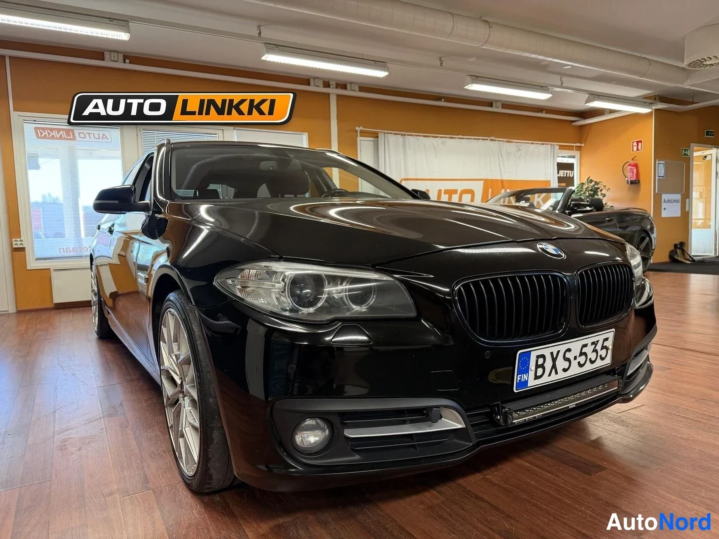 Etuviisto, nokka oikealle: Etukulmakuva mustasta BMW-autosta sisätiloissa, keulapaneeli ja osa kyljestä näkyvissä, rekisterikilpi BX-535.