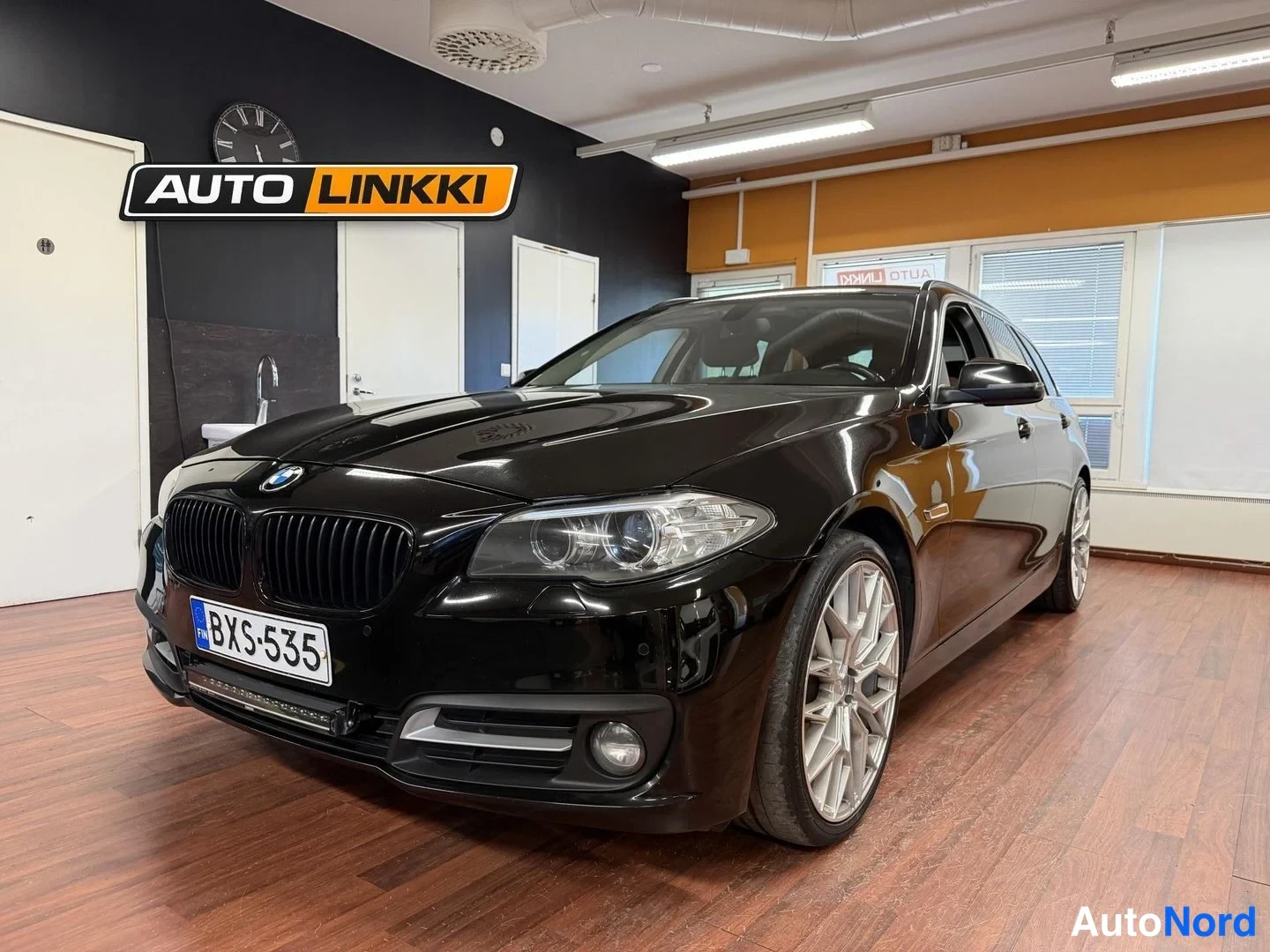 Etuviisto, nokka vasemmalle: Musta BMW-station wagon etuviistosta vasemmalta sisätiloissa; etupaneeli ja kylki näkyvät, kromilattialla hopeiset vanteet, kilpi BX-535.