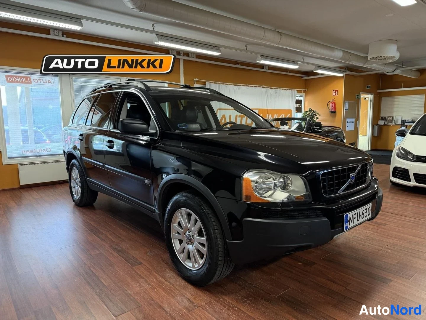 Etuviisto, nokka oikealle: Musta Volvo XC90 -maastauto etukulmasta autosalissa, rekisterikilpi.