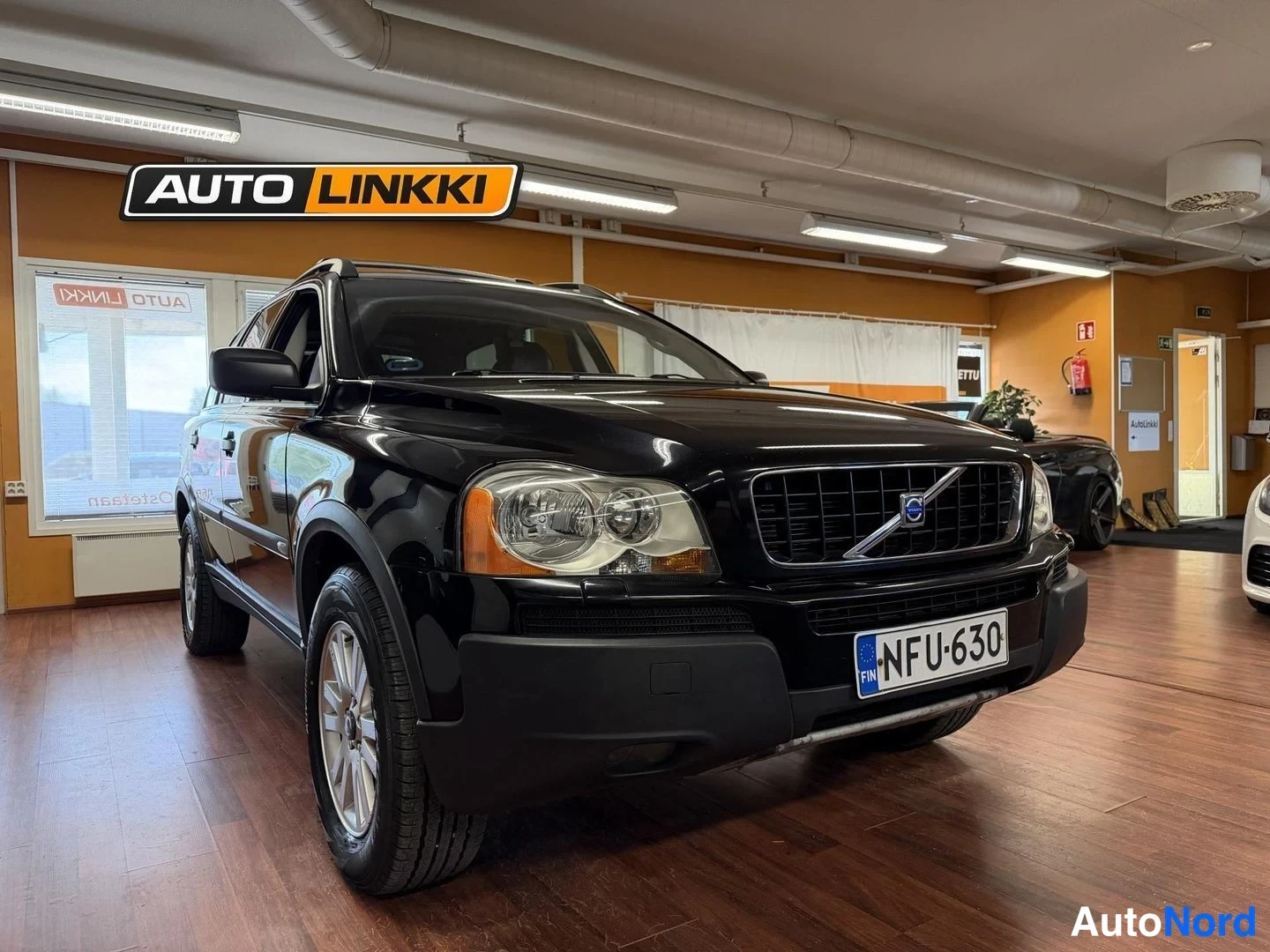 Etuviisto, nokka oikealle: Musta Volvo XC90 etukulmasta sisätiloissa, keulakoriste ja rekisterikilpi näkyvissä.