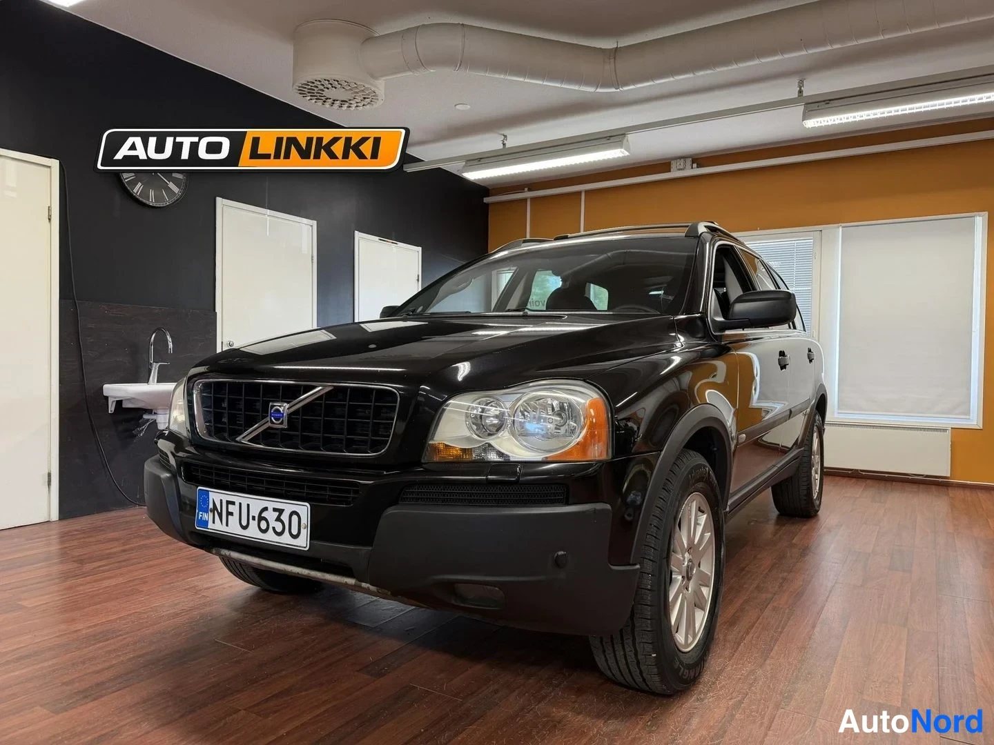 Etuviisto, nokka vasemmalle: Musta Volvo XC90 etukulmasta vasemmalle käännettynä huoneessa, rekisterikilpi.
