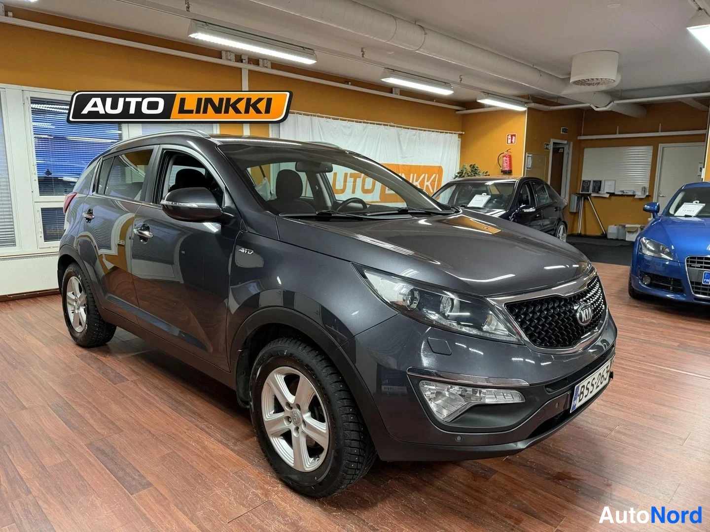 Etuviisto, nokka oikealle: Kia Sportage -maastauto tummanharmaana AUTO LINKKI -tilan lattiassa, etukulmanäkyvasemmalta
