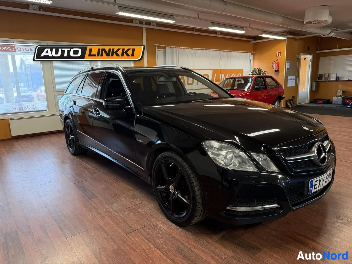 Etuviisto, nokka oikealle: Musta Mercedes-Benz farmarivalauto esillä sisätiloissa, etukulma oikealta; tummat kevytmetallivanteet ja etupuskurin anturit näkyvät.
