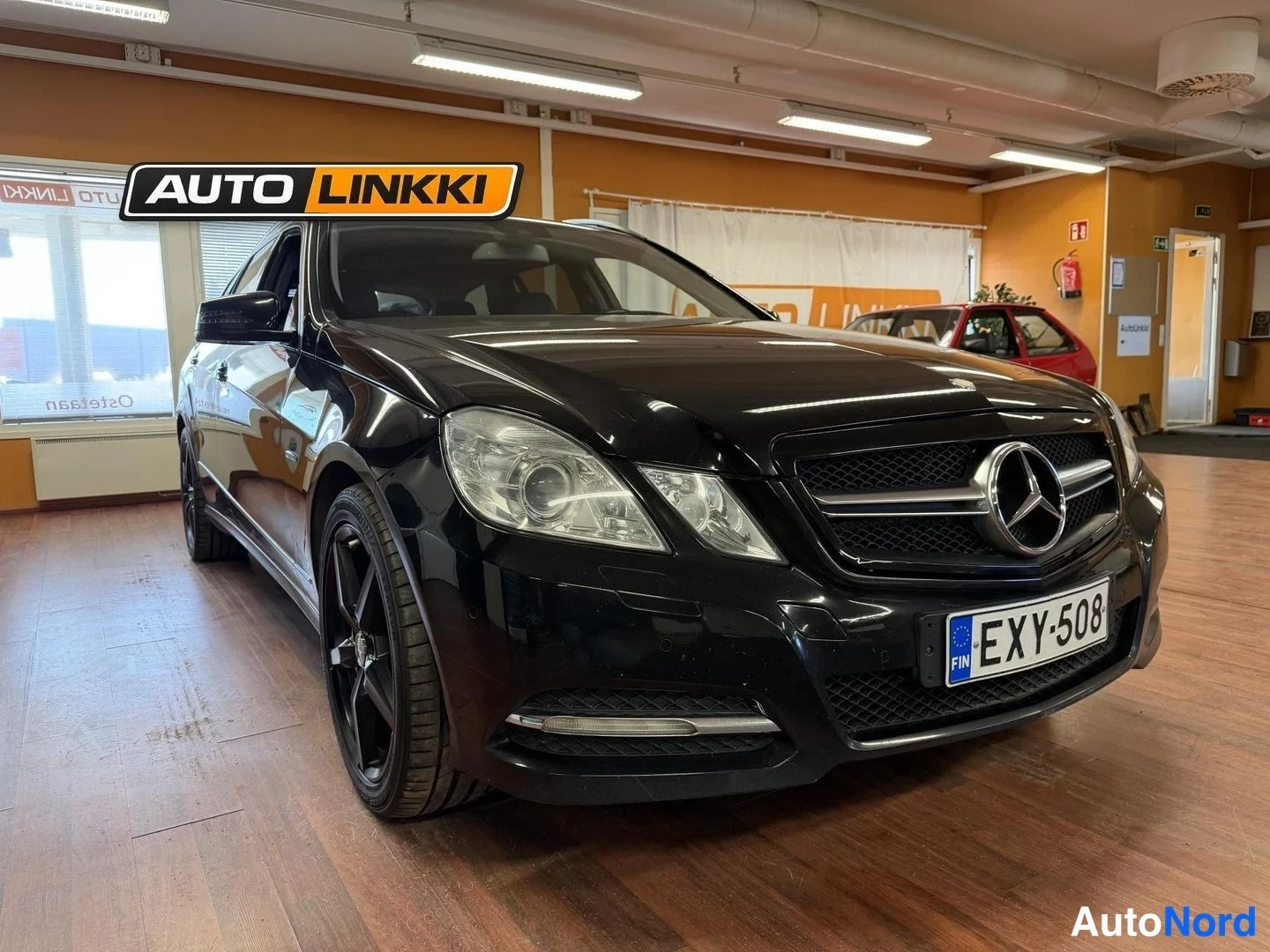 Etuviisto, nokka oikealle: Musta Mercedes-Benz farmari sisätiloissa etukulmasta oikealta; rekisterikilpi ja tummat kevytmetallivanteet näkyvät.