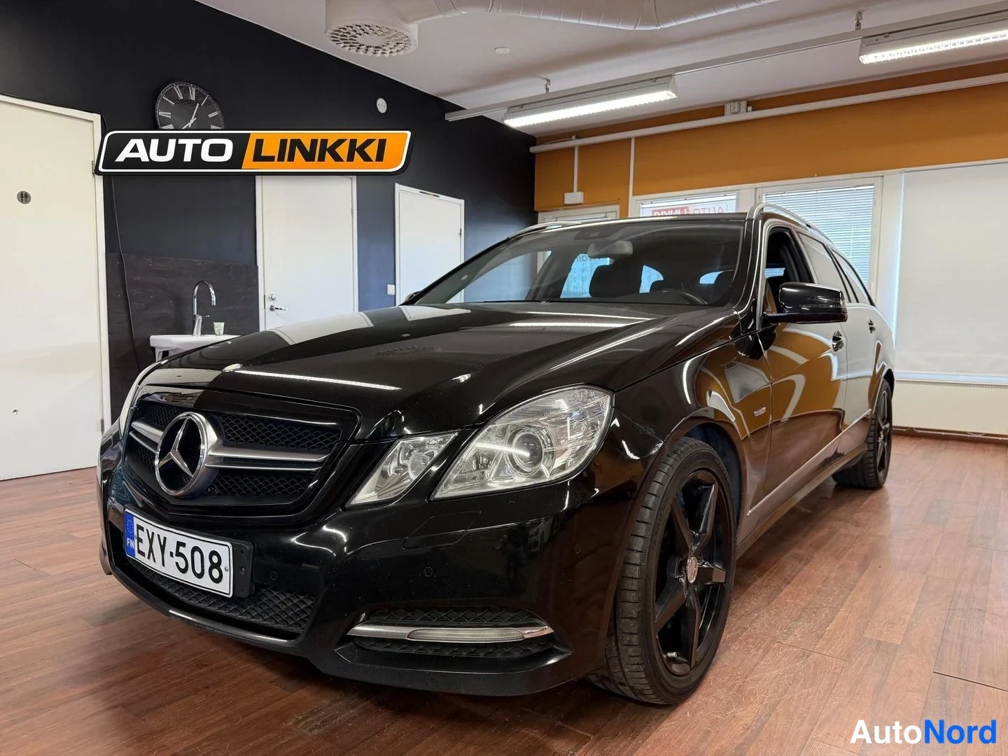 Etuviisto, nokka vasemmalle: Musta Mercedes-Benz farmari sisätiloissa etukulmasta vasemmalta; rekisterikilpi ja mustat kevytmetallivanteet näkyvät.